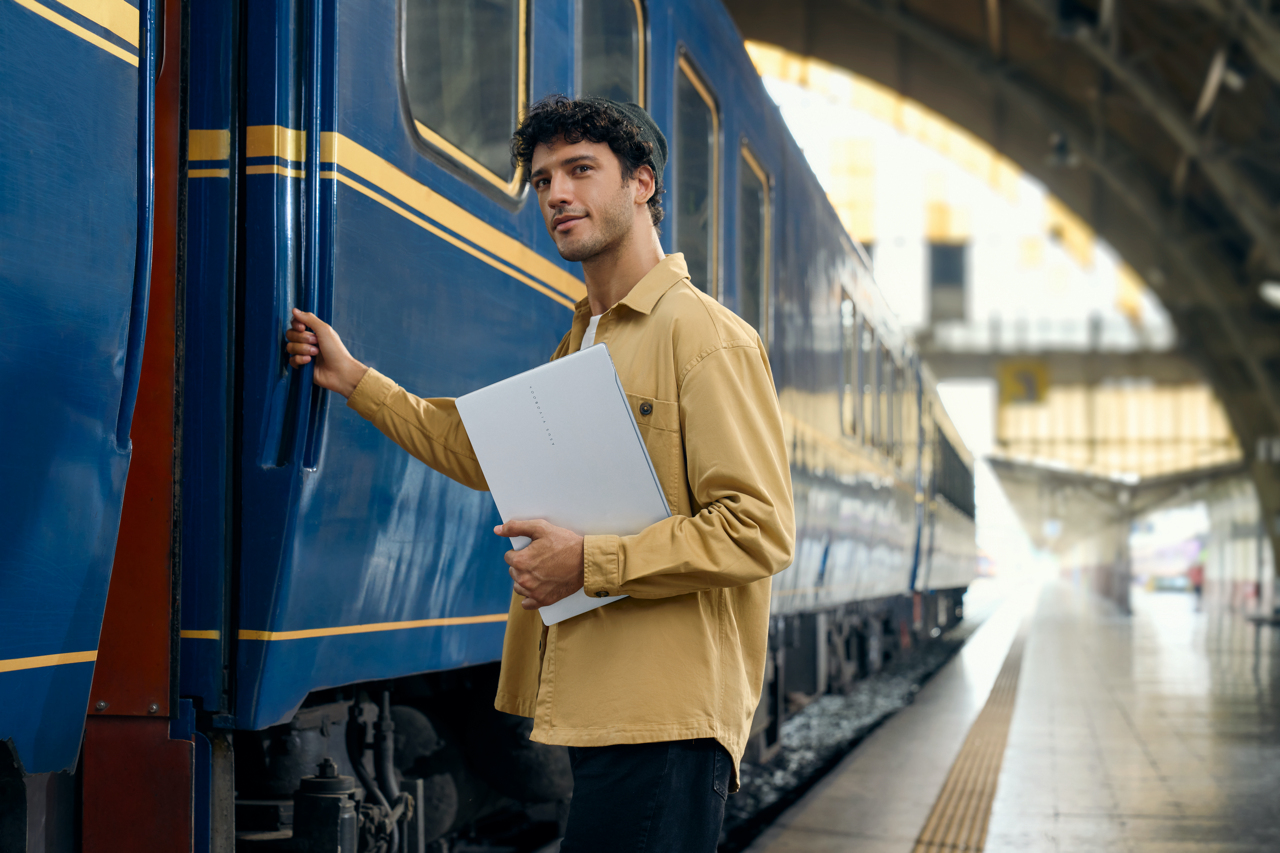 Mann mit Laptop an einem blau-gelben Zug. Der Mann trägt eine beige Jacke.