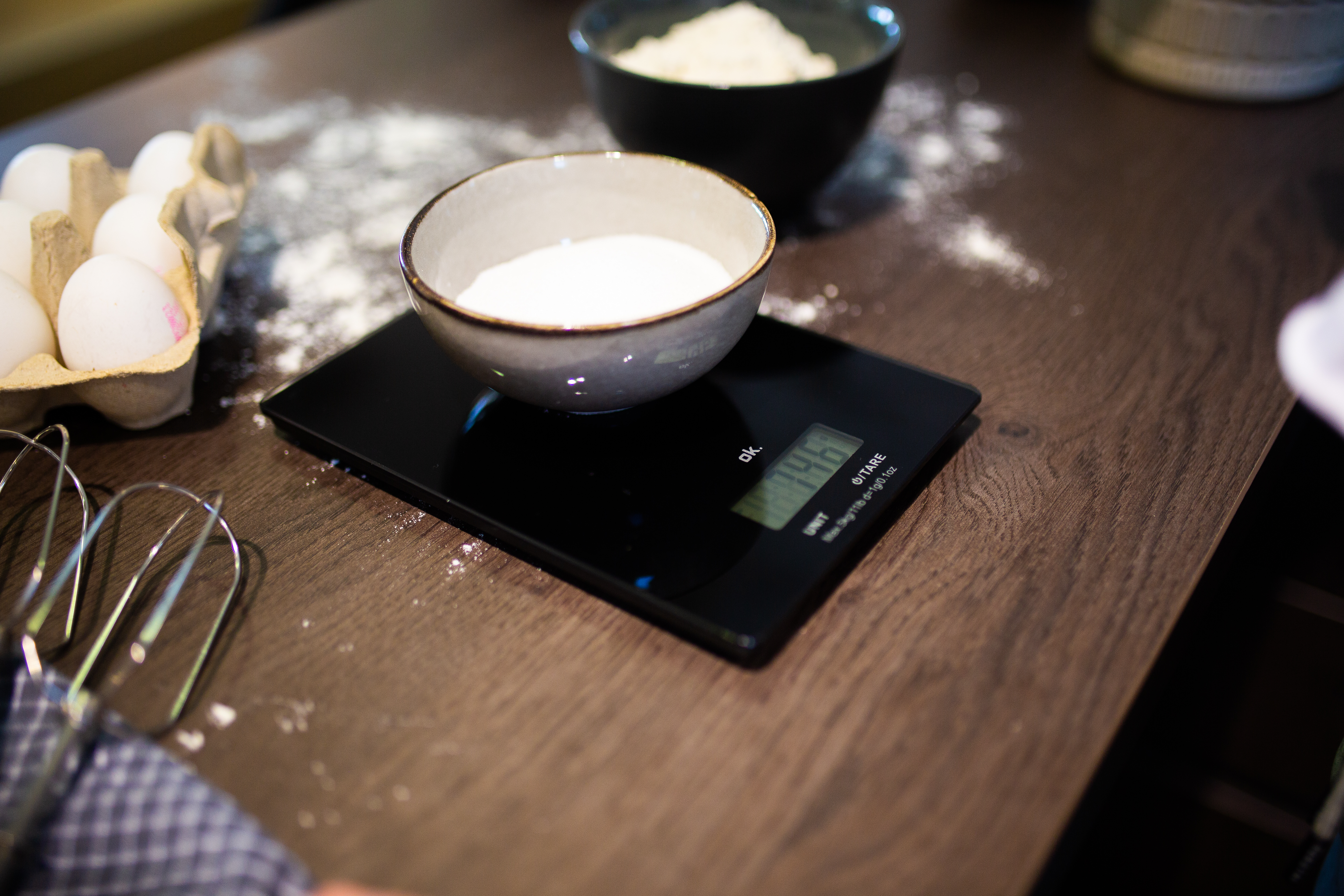 Waga z miską cukru. Jajka, mąka i trzepaczka w tle.