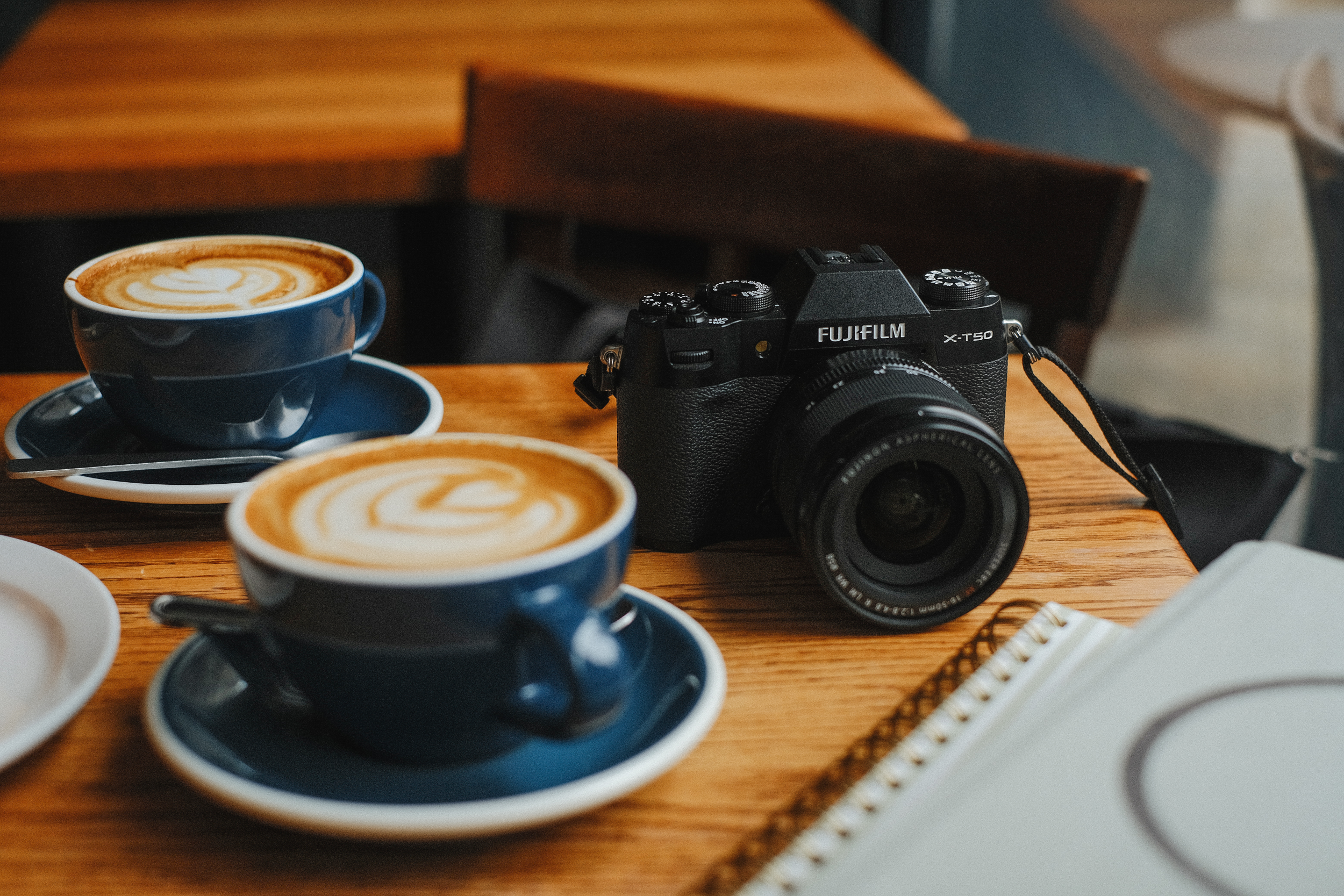 Zwei dunkelblaue Kaffeetassen mit Latte Art und eine Kamera auf einem Holztisch.
