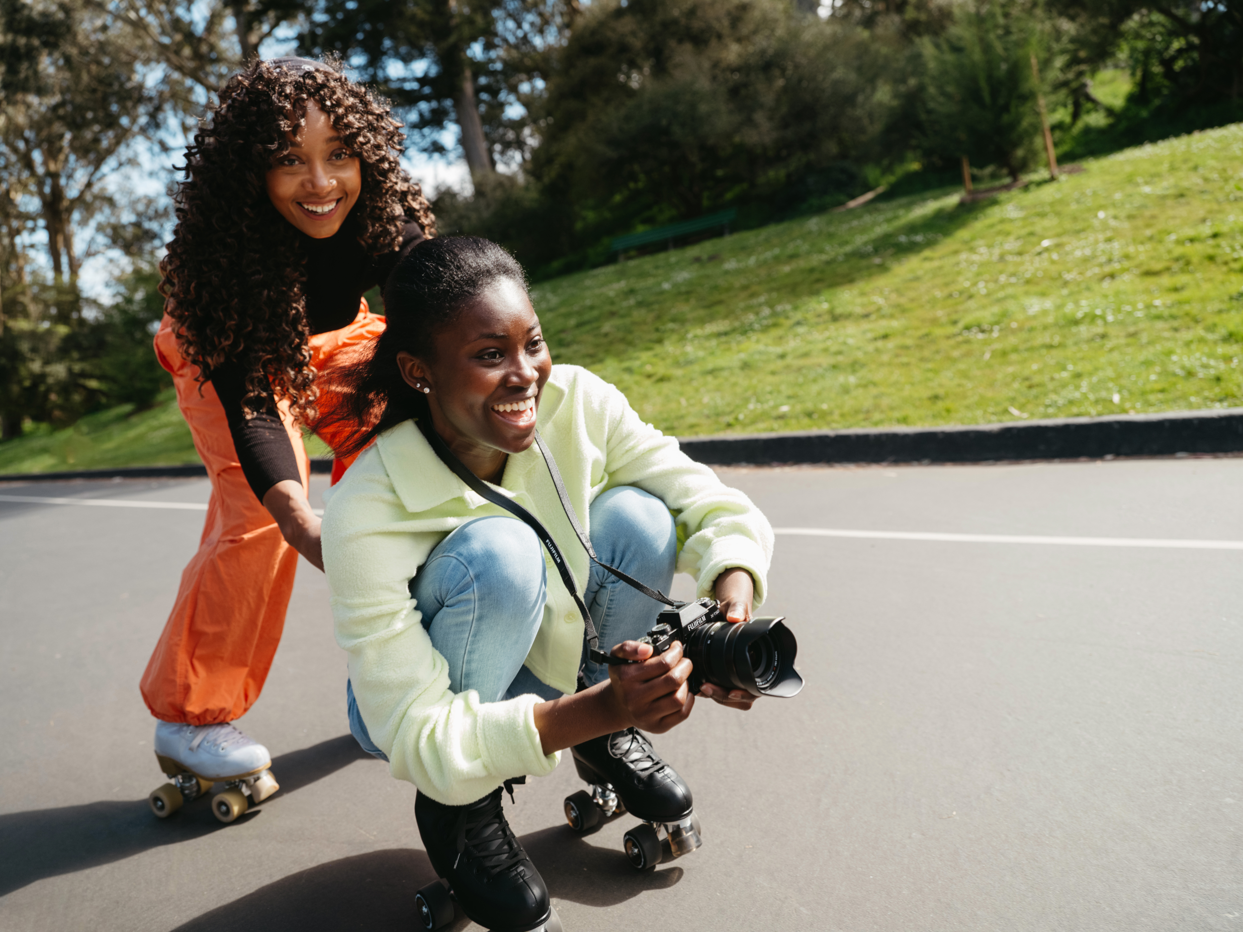 Zwei Frauen auf Rollschuhen, eine macht Fotos. Grüner Hintergrund und Asphaltstraße.