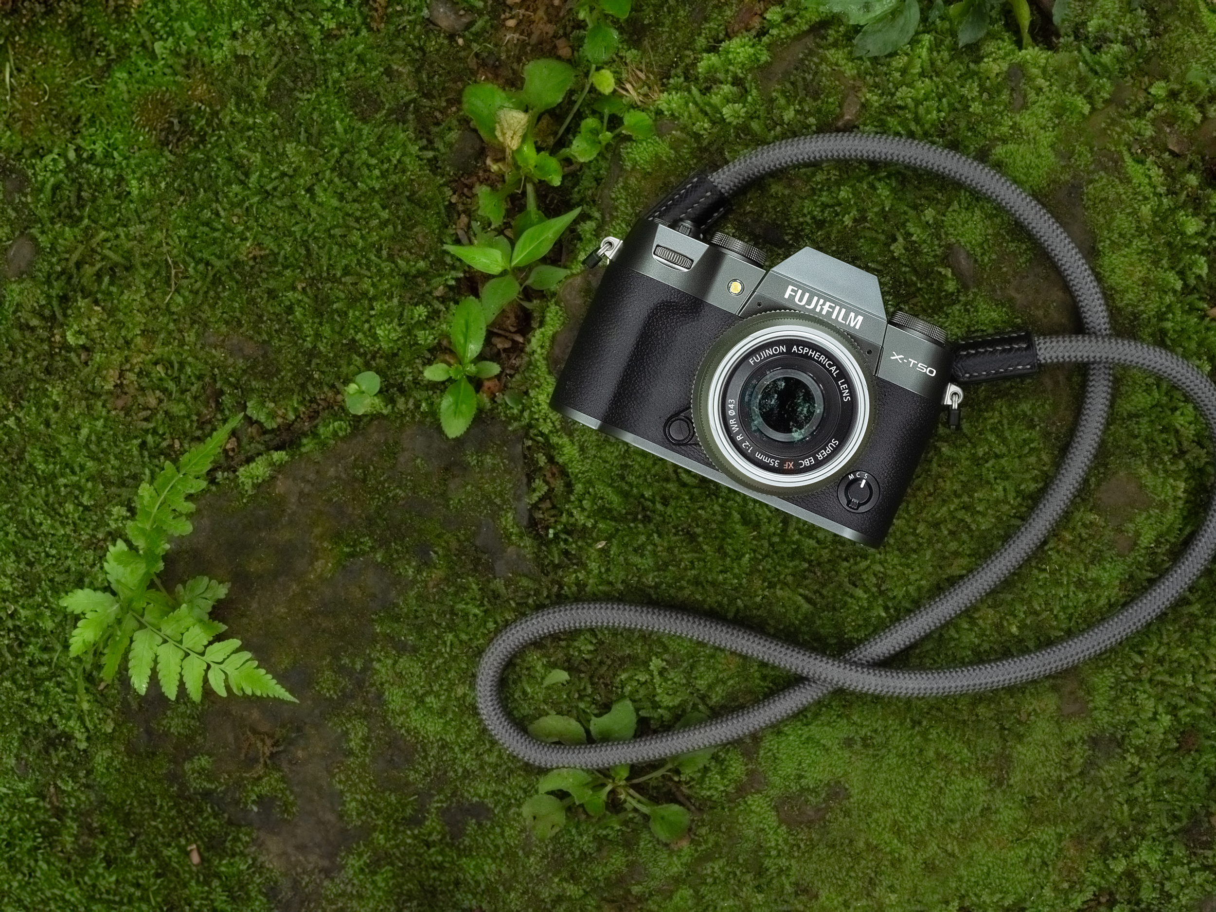Eine Person hält eine Fujifilm X-T50 Kamera. Eine andere Person sitzt im Hintergrund. Die Kamera hat ein Objektiv.