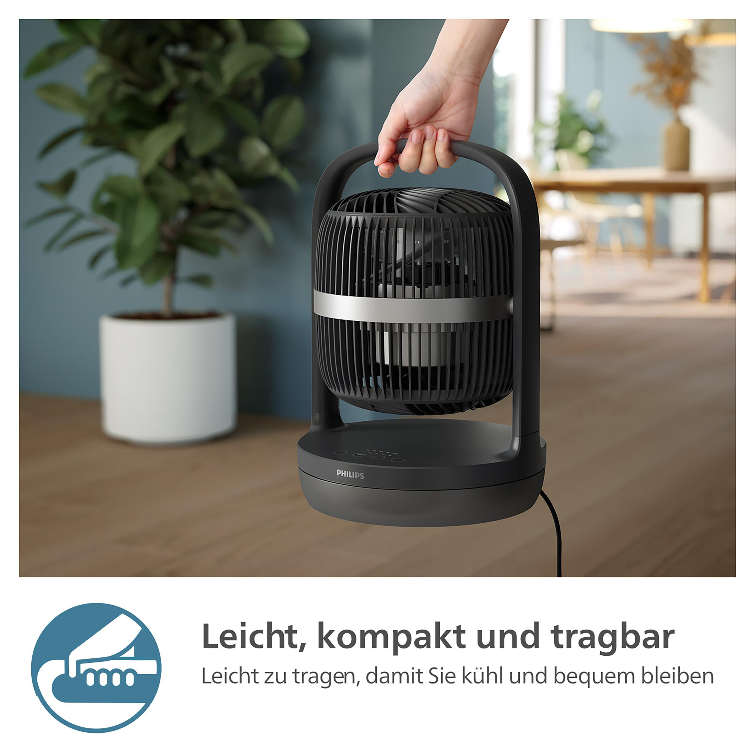 Hand hält einen schwarzen Ventilator, Blumentopf im Hintergrund.