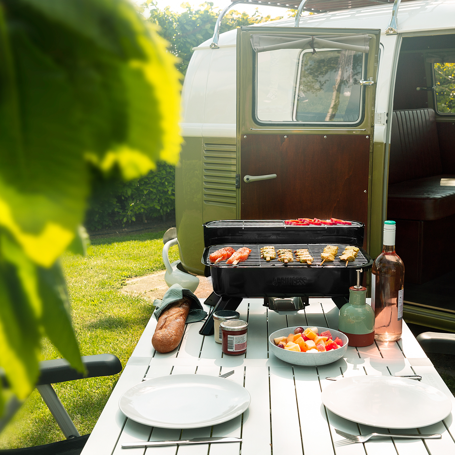 Tavolo allestito per un barbecue. Include grill, cibo, piatti, bottiglie e un furgone verde sullo sfondo.