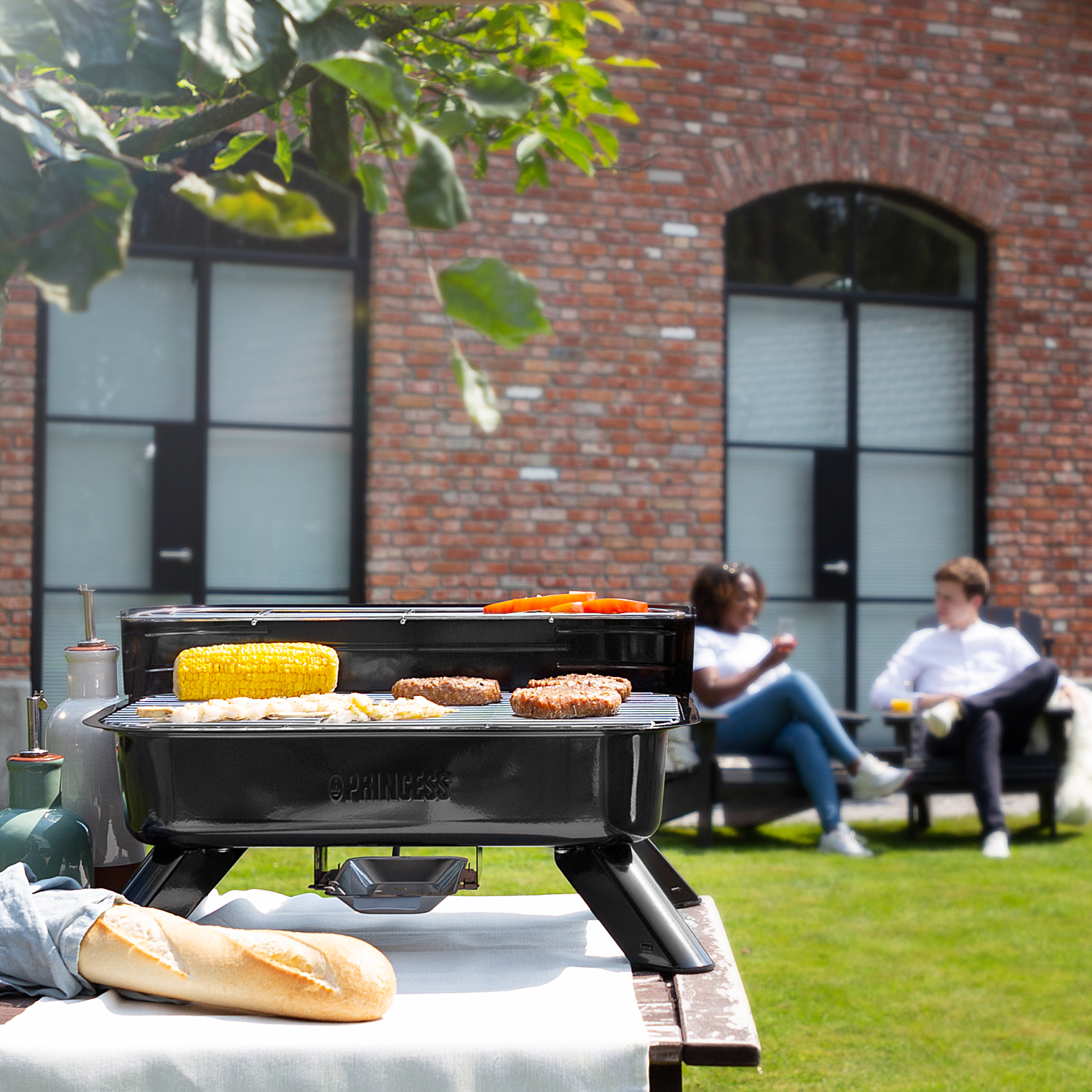 Un barbecue con cibo sopra, una baguette e due persone che si rilassano sullo sfondo.