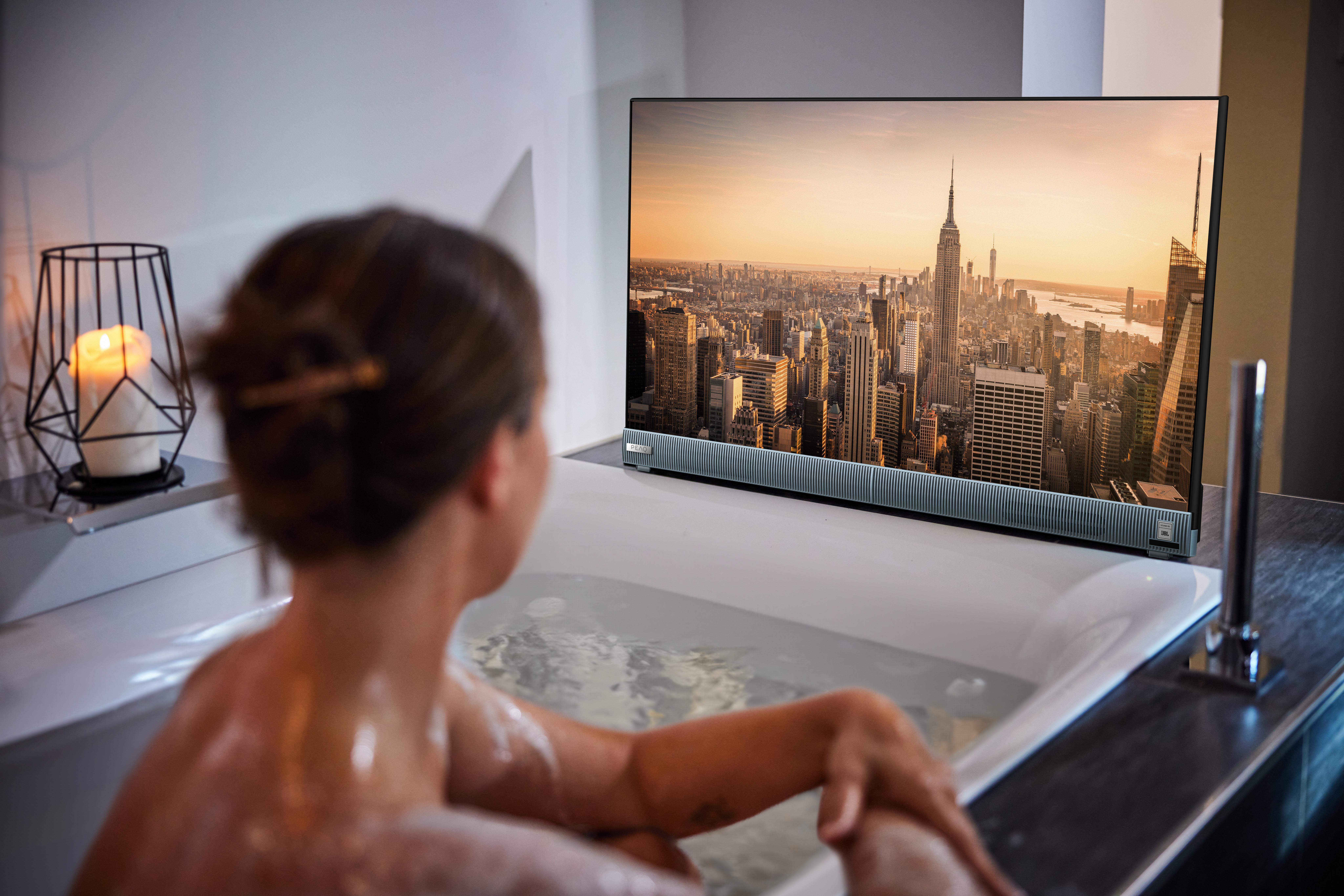 Frau in Badewanne sieht TV mit NYC Skyline; Kerze und Deko daneben.