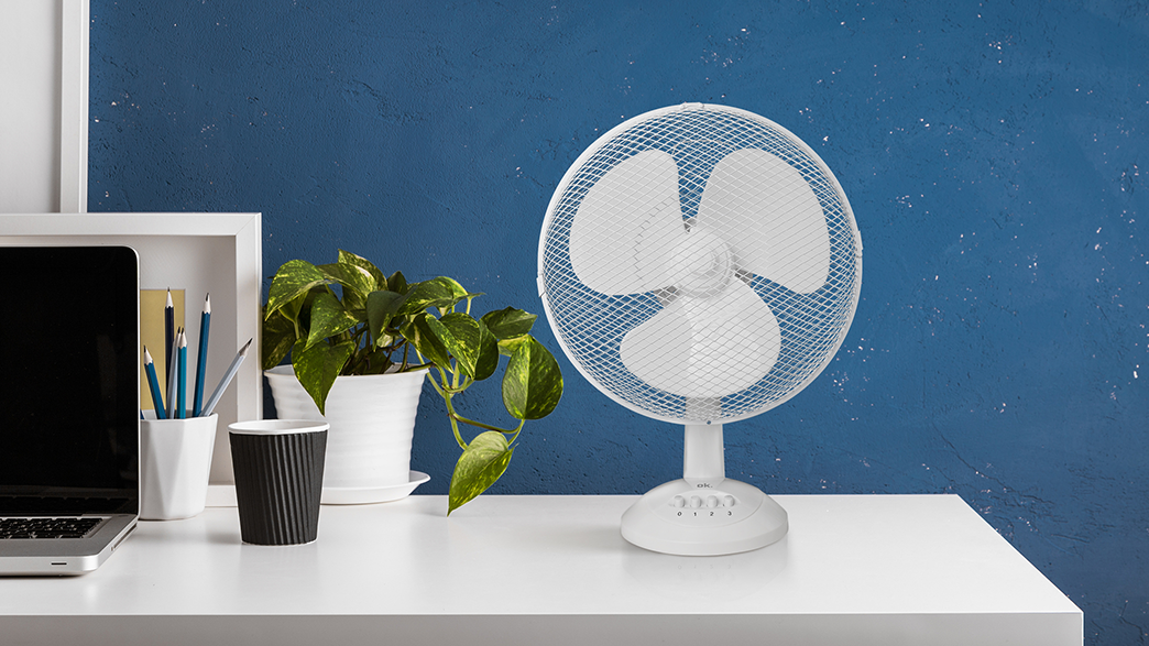 Fehér asztal ventilátorral, laptop, ceruzák és egy cserepes növény kék fal előtt.