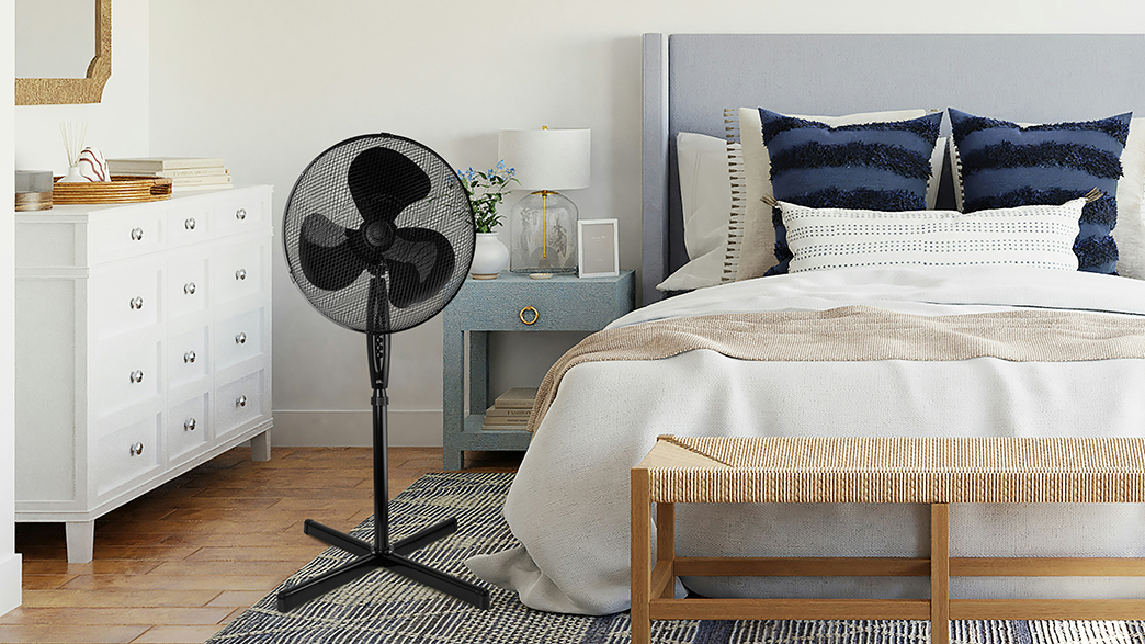 Fekete álló ventilátor egy hálószobában ággyal, éjjeliszekrénnyel és szőnyeggel.