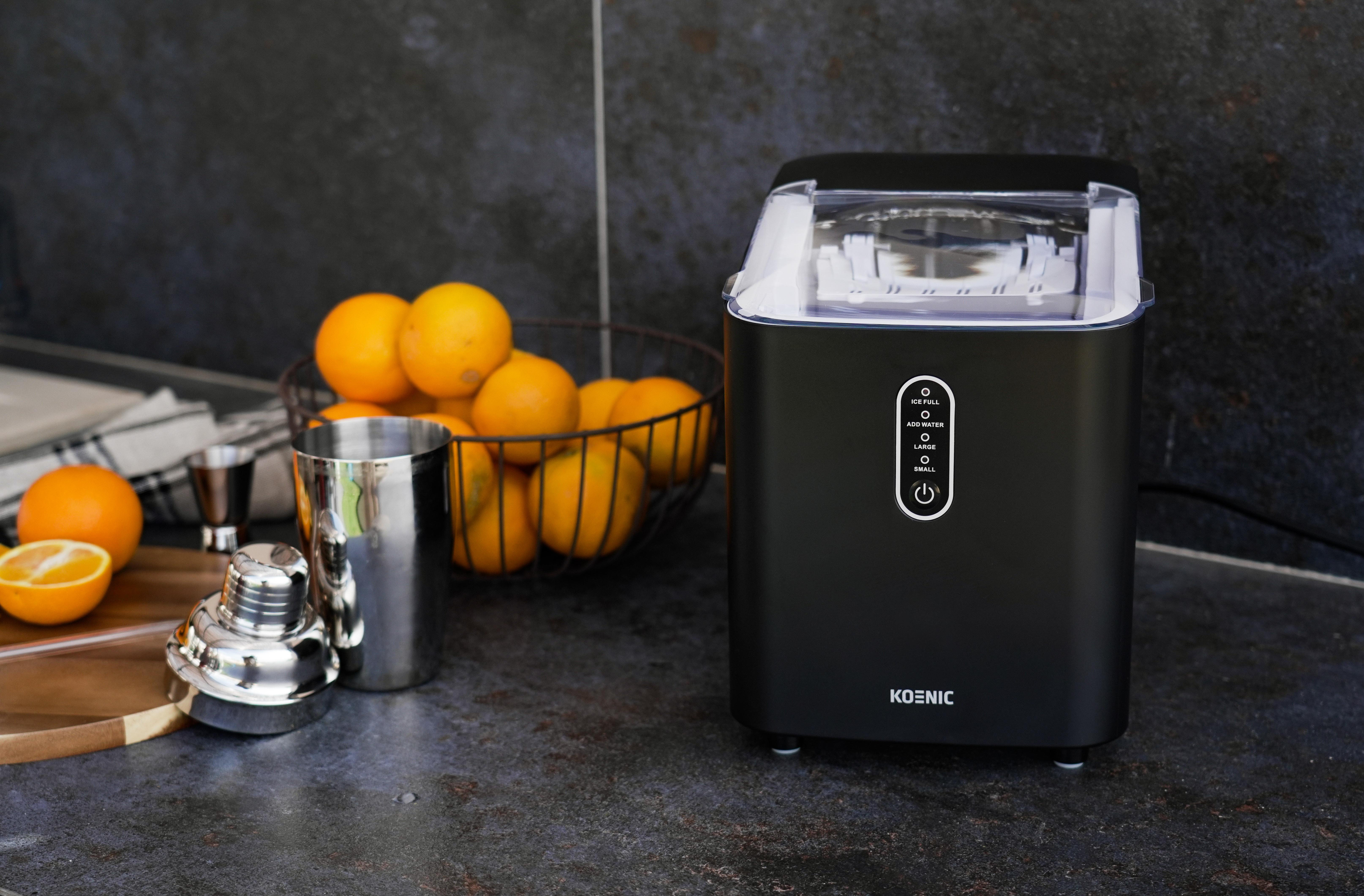 Schwarzer Eisbereiter auf einer Theke mit Orangen, einem Shaker und einem Messbecher. Dunkler Fliesenhintergrund.