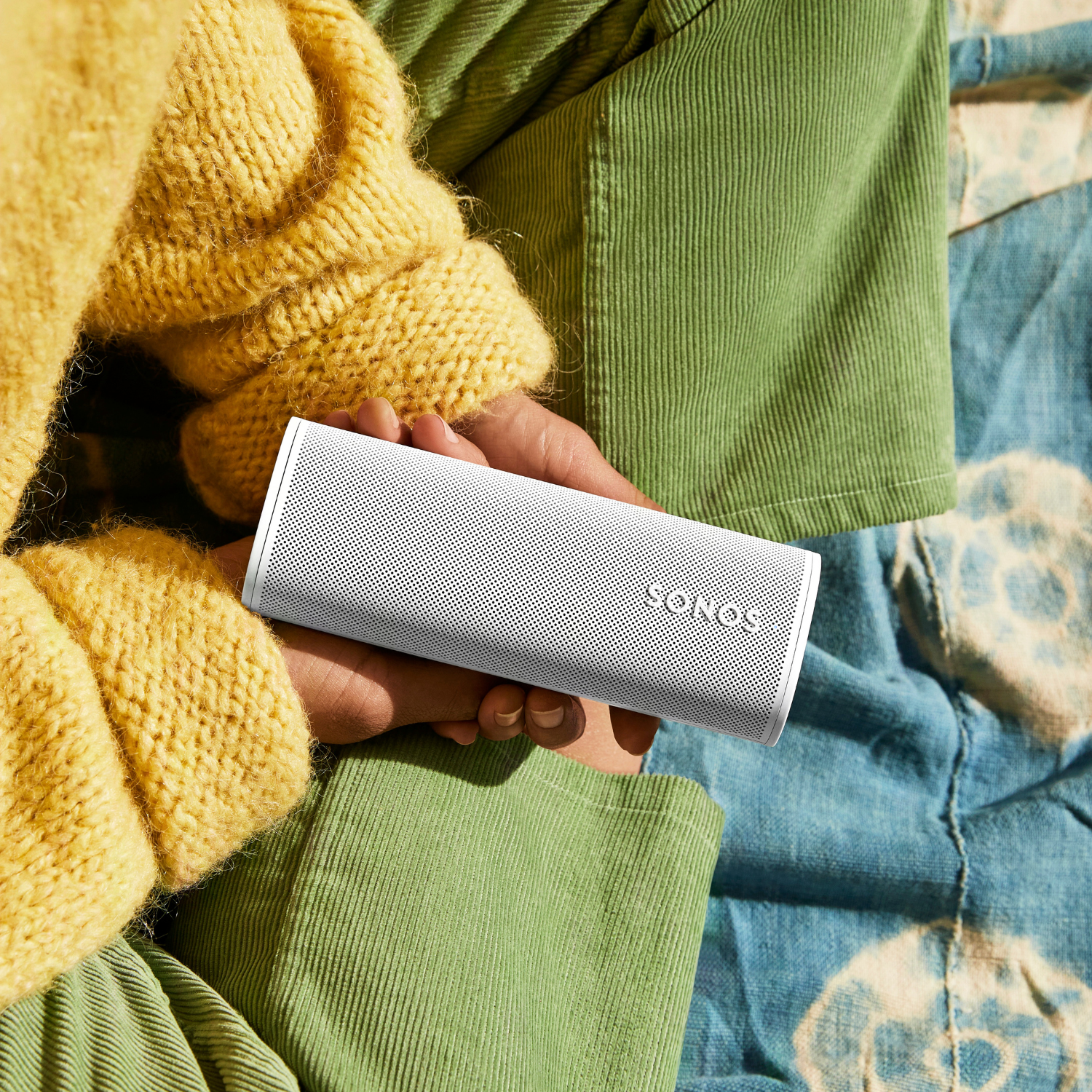 Persona sosteniendo un altavoz Sonos blanco; lleva un suéter amarillo y pantalones verdes.