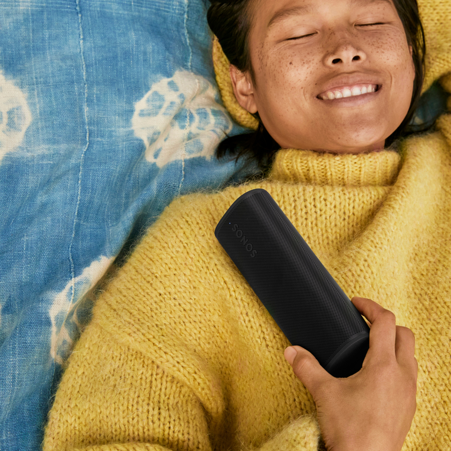 Una mujer sosteniendo un altavoz Sonos negro. Está acostada en una manta azul.