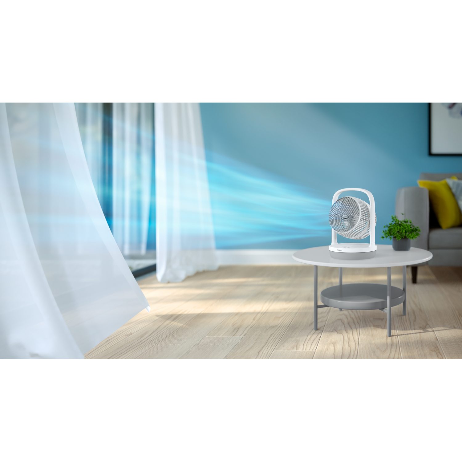 A white fan on a table blowing air toward a blue wall. Curtains are on the left.