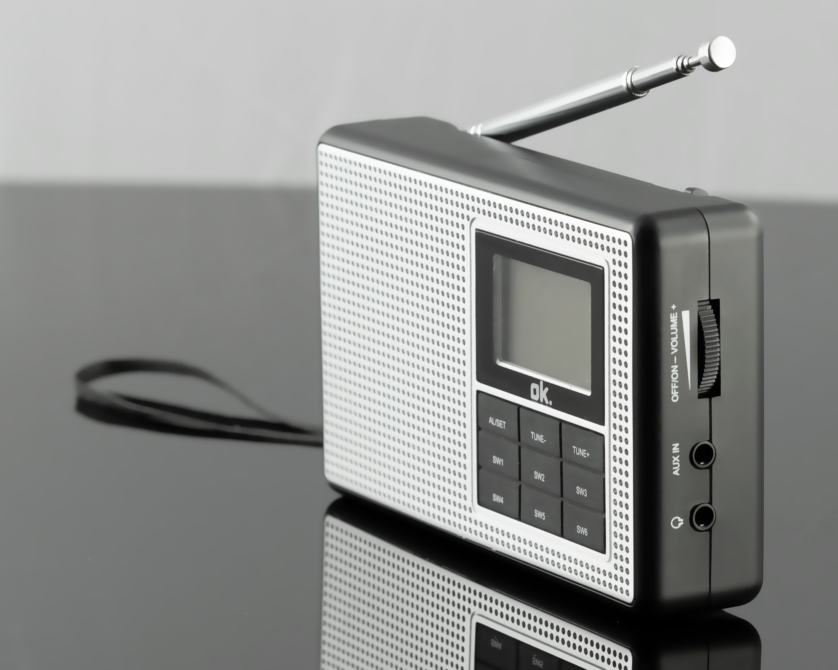 Srebrno-czarne radio z anteną, ekranem, przyciskami i wejściem aux. Odbicie na czarnej powierzchni.