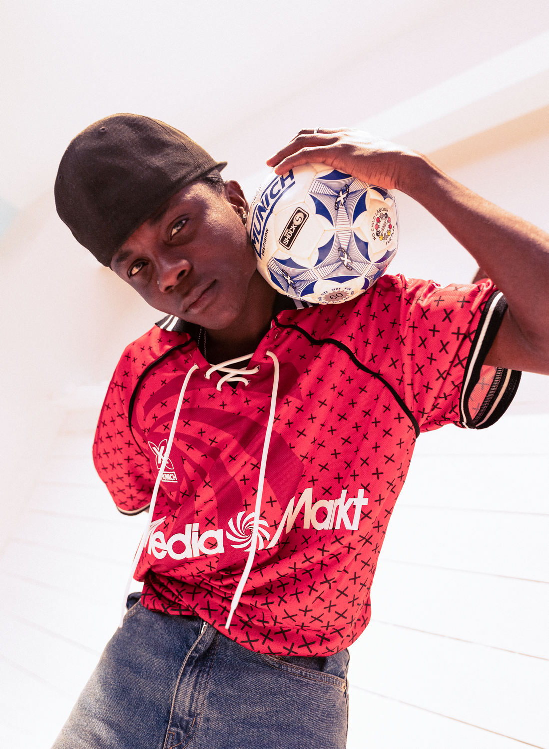 Un joven con gorra negra sostiene un balón de fútbol, vistiendo una camiseta roja con cruces negras.