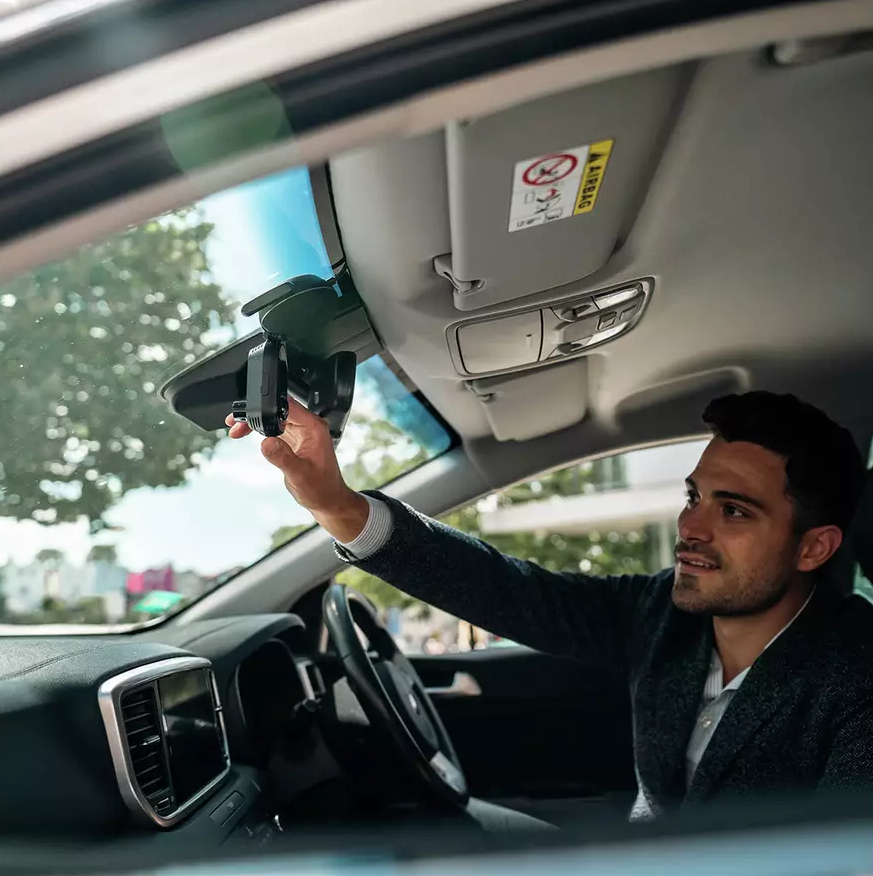 Mężczyzna w samochodzie reguluje uchwyt na telefon na lusterku wstecznym, widoczny przez przednią szybę.