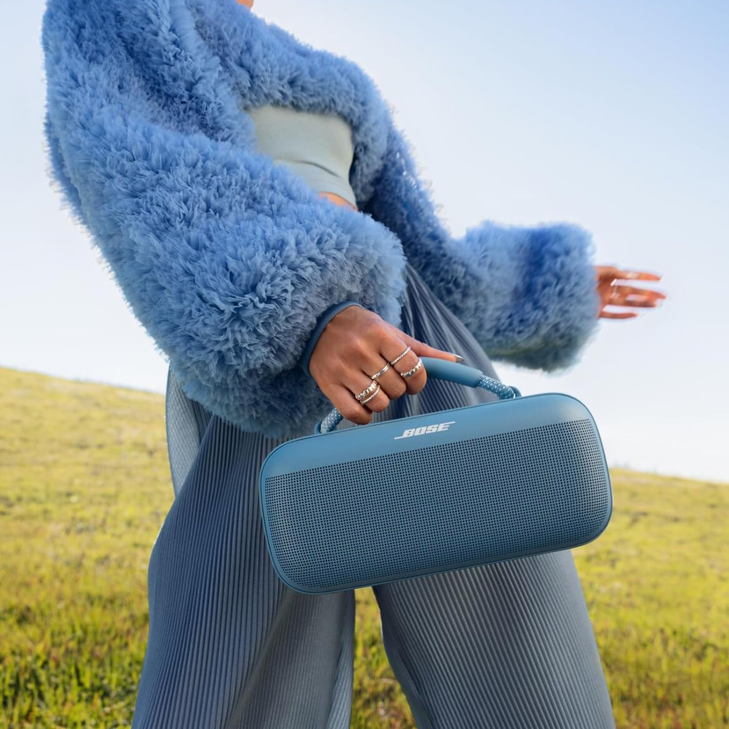 Persona sosteniendo un altavoz Bose azul. Chaqueta de piel azul. Pantalones plisados azules. Fondo de hierba verde.