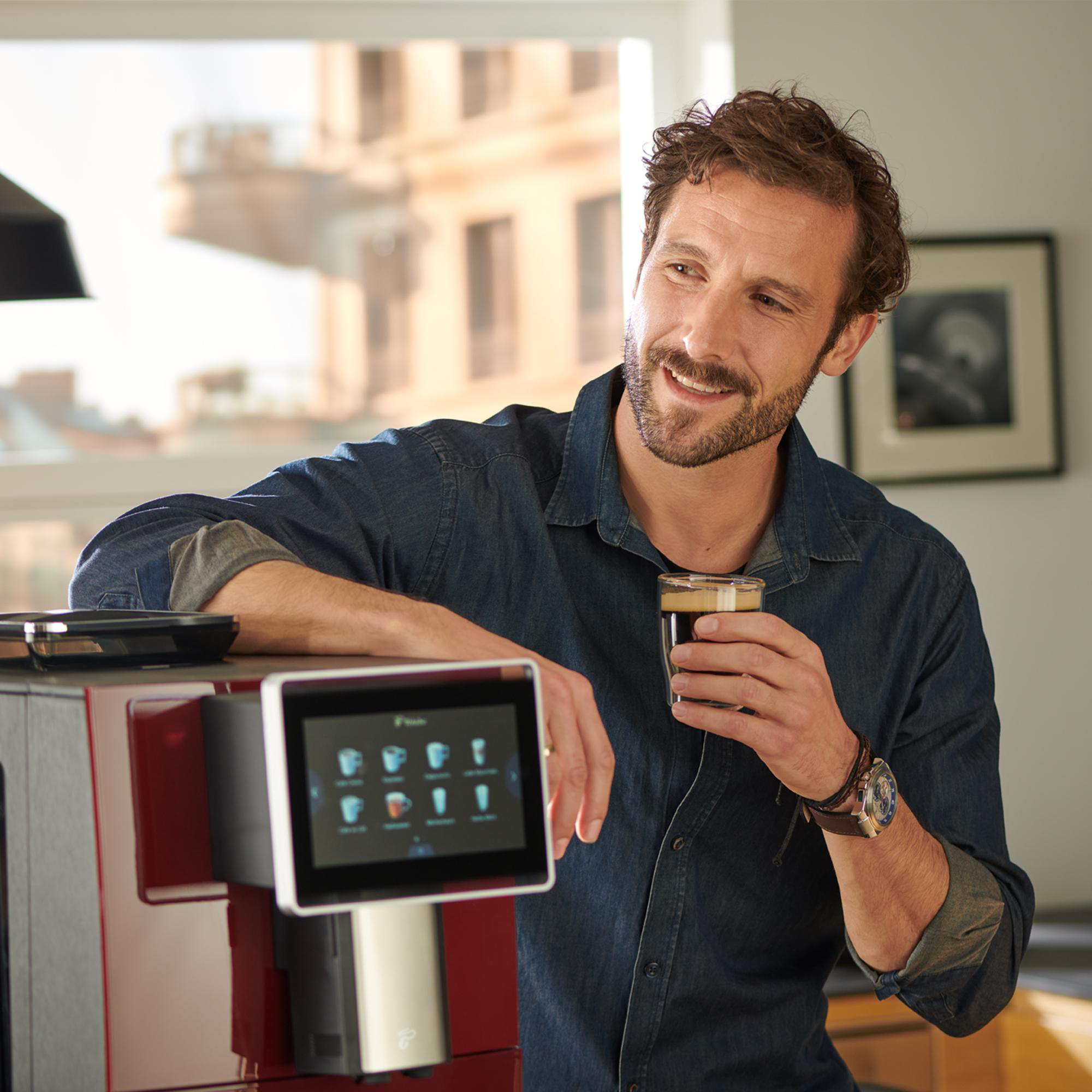 Ein Mann hält ein Kaffeeglas in der Nähe einer Kaffeemaschine und lächelt.