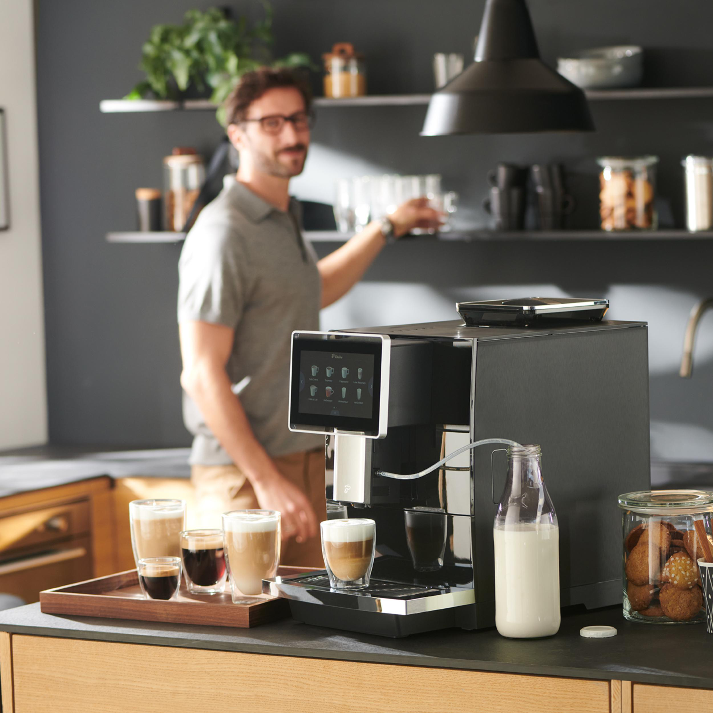 Küche mit Kaffeemaschine, Kaffeegläsern, Milchflasche und einem Mann, der nach einem Regal greift.