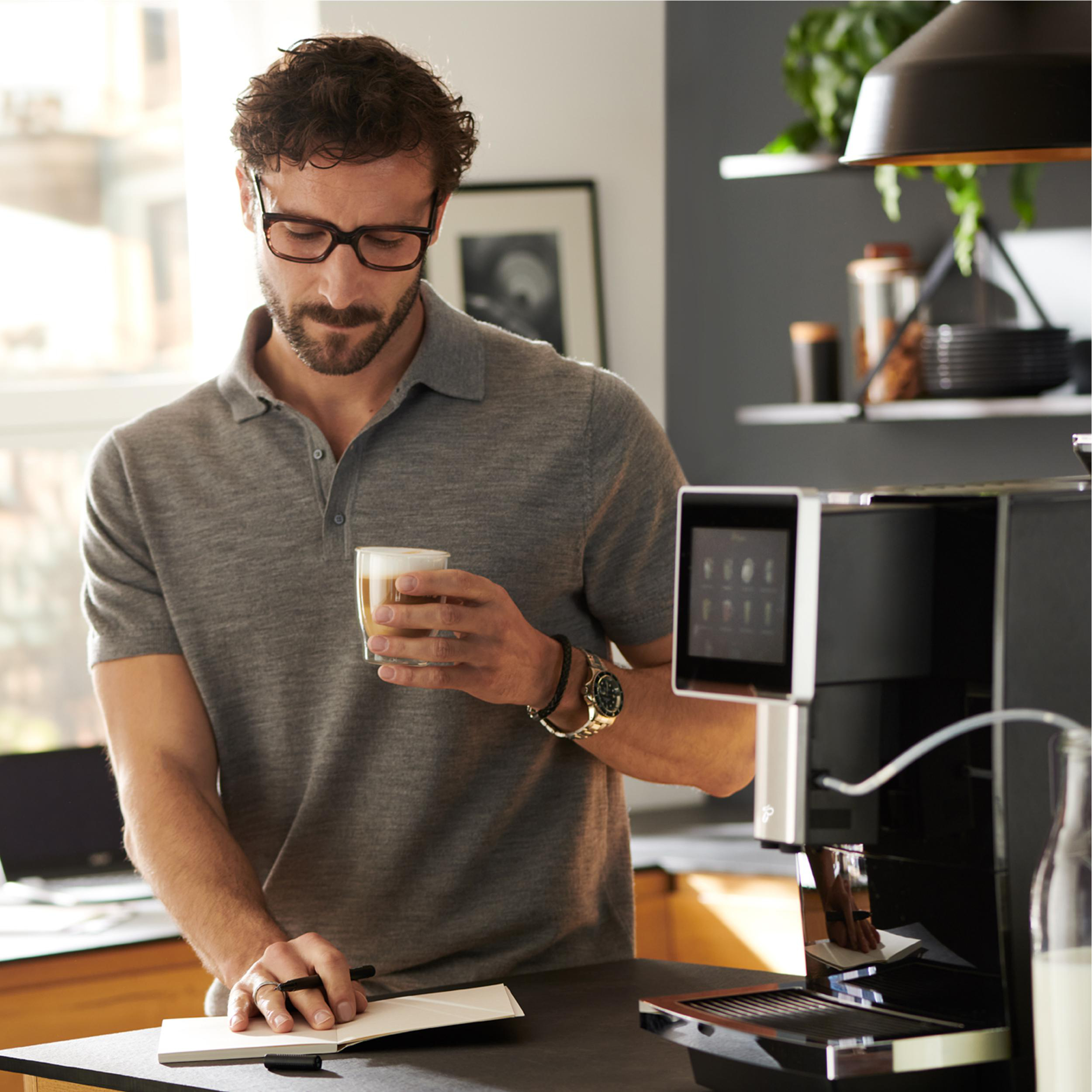Ein Mann hält eine Kaffeetasse, mit Kaffeemaschine und Notizbuch.