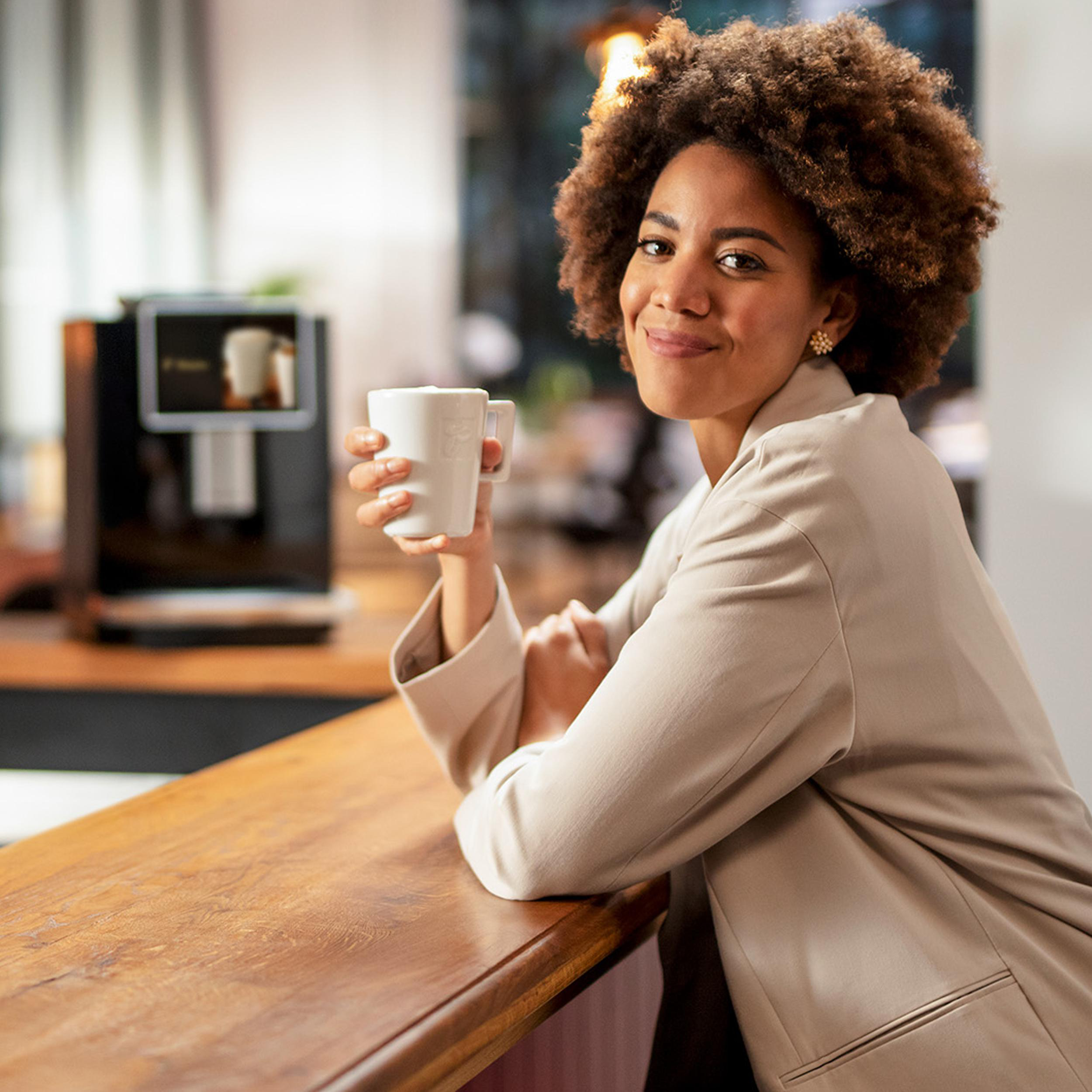 Eine lächelnde Frau hält eine weiße Tasse in der Nähe einer Holztheke, im Hintergrund eine Kaffeemaschine.