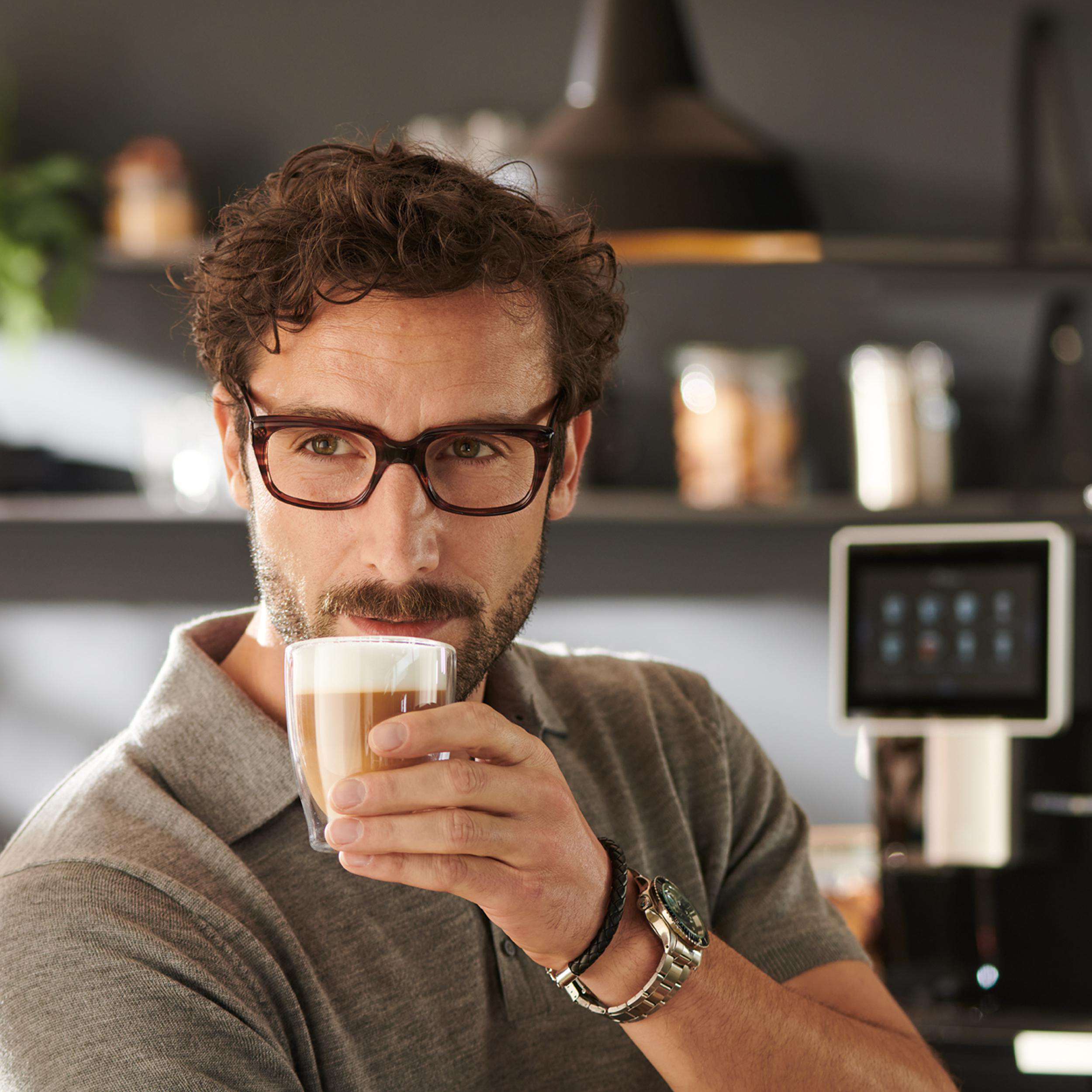 Mann trinkt einen Latte Macchiato. Er trägt eine Brille und eine Uhr und befindet sich in einer Küche.