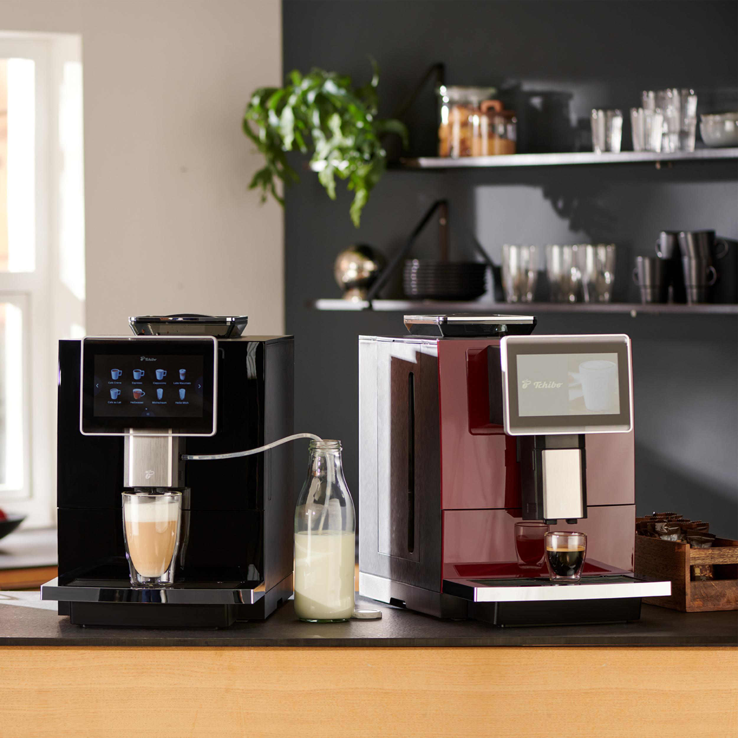 Zwei schwarz-rote Kaffeemaschinen mit Touchscreens auf einer hölzernen Arbeitsplatte, neben Milch.