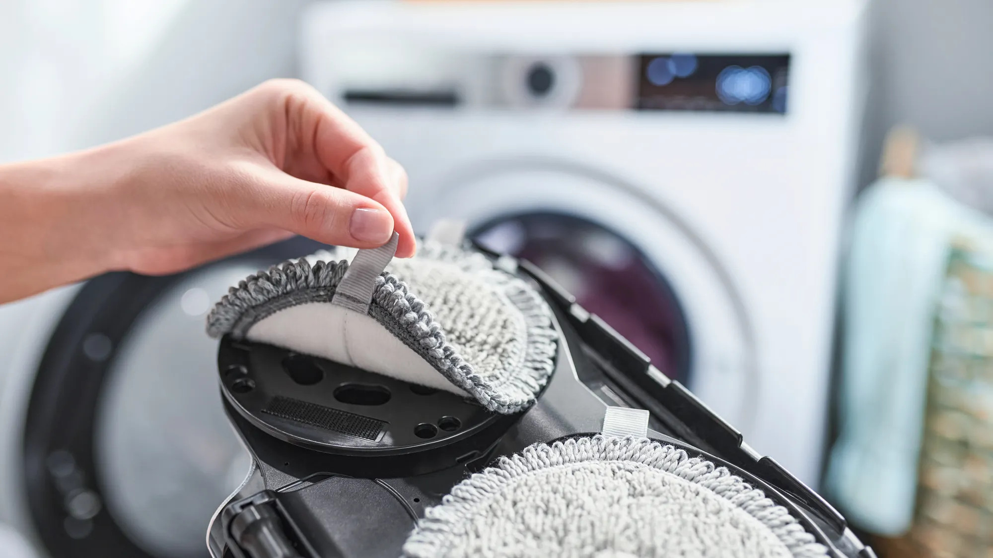 Ręka wyciąga nakładkę z robota sprzątającego. W tle pralka.