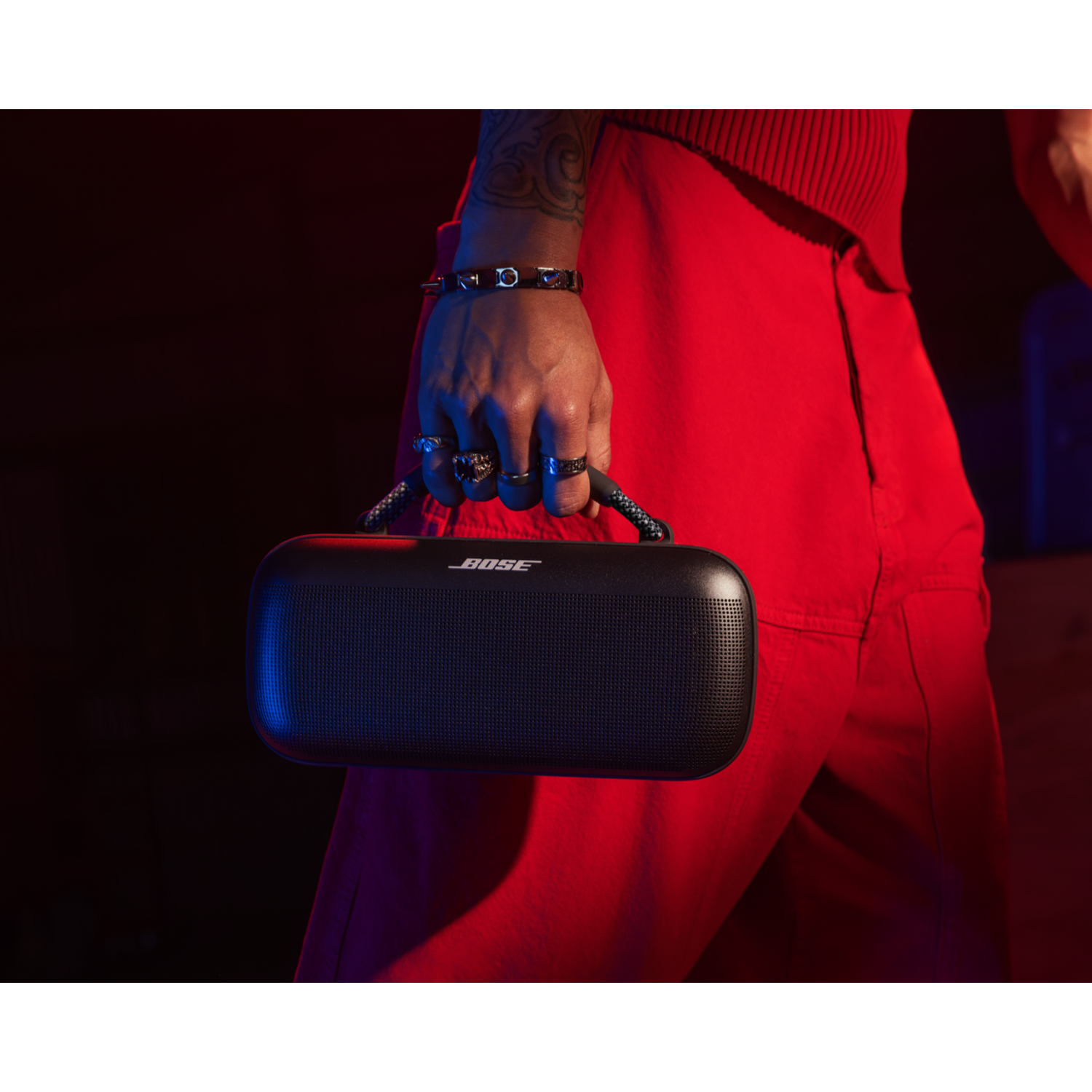 Person holding a black Bose speaker. They are wearing red pants and holding the speaker with a hand.