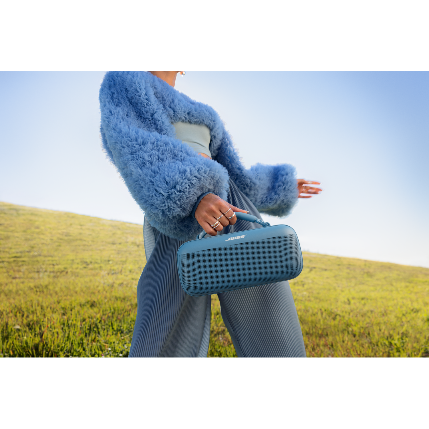 Een vrouw in een blauwe bontjas die een blauwe Bose-luidspreker vasthoudt op gras in de buitenlucht, met een blauwe lucht op de achtergrond.