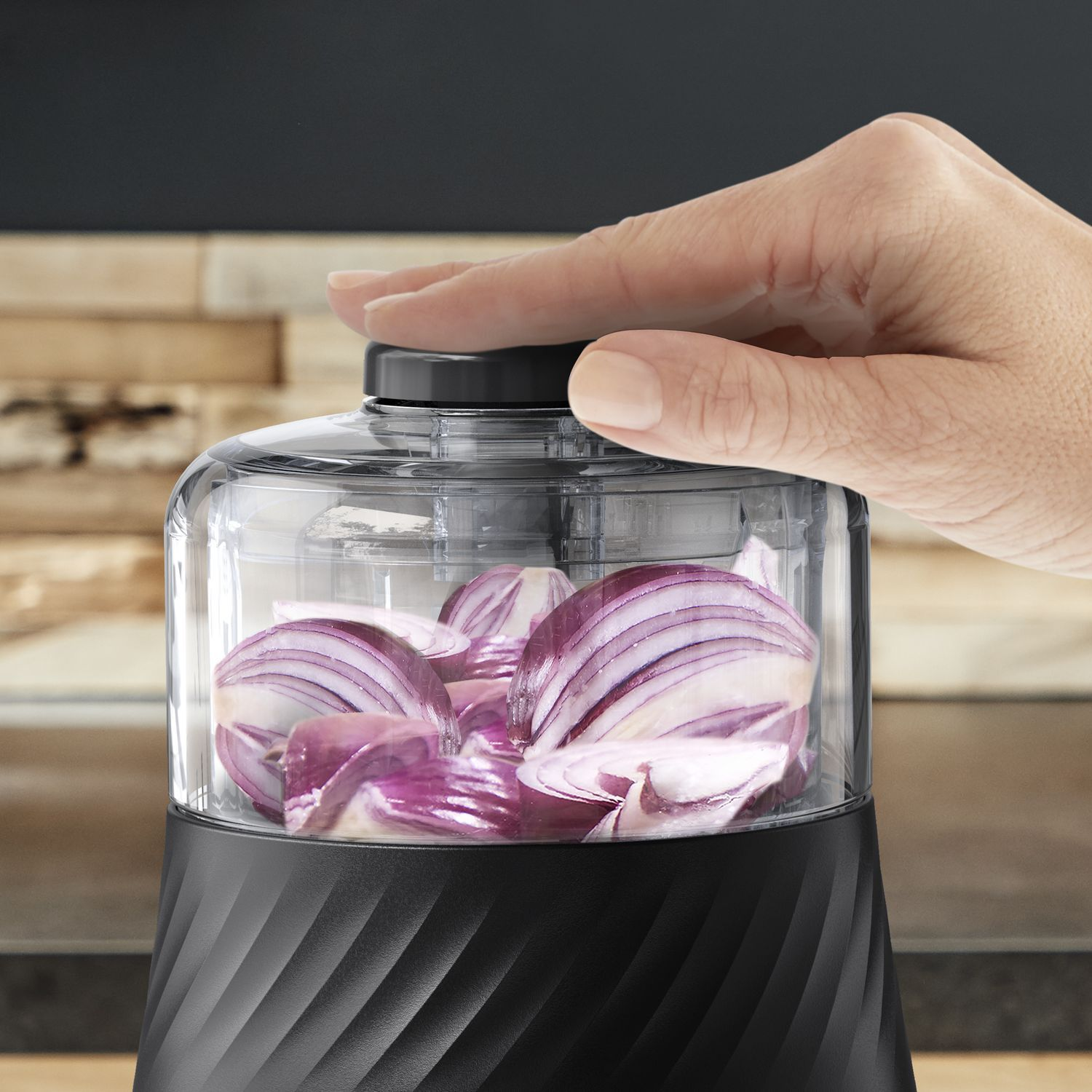 Hand presses a top button of a black and clear food chopper. Sliced red onions inside the container.