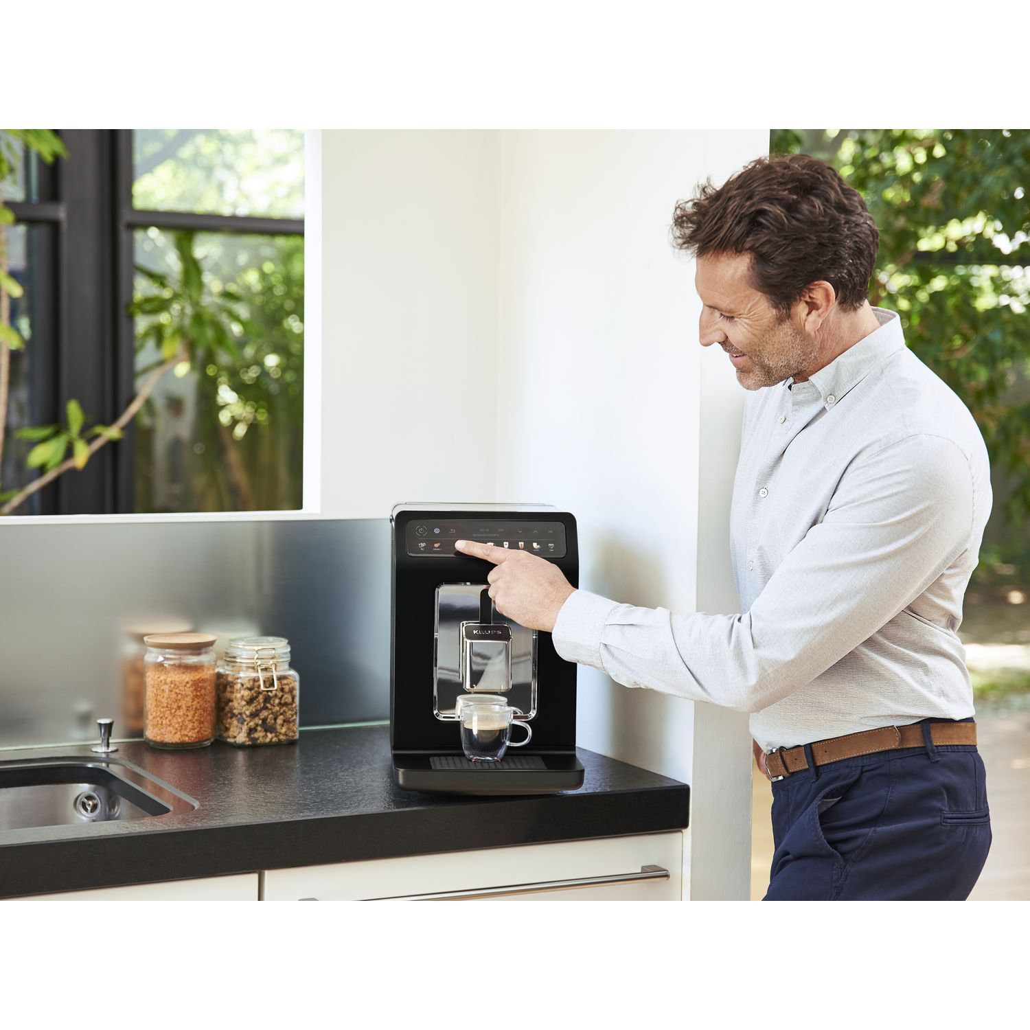 A man using a black coffee machine with a glass cup, kitchen counter and jars in the back.