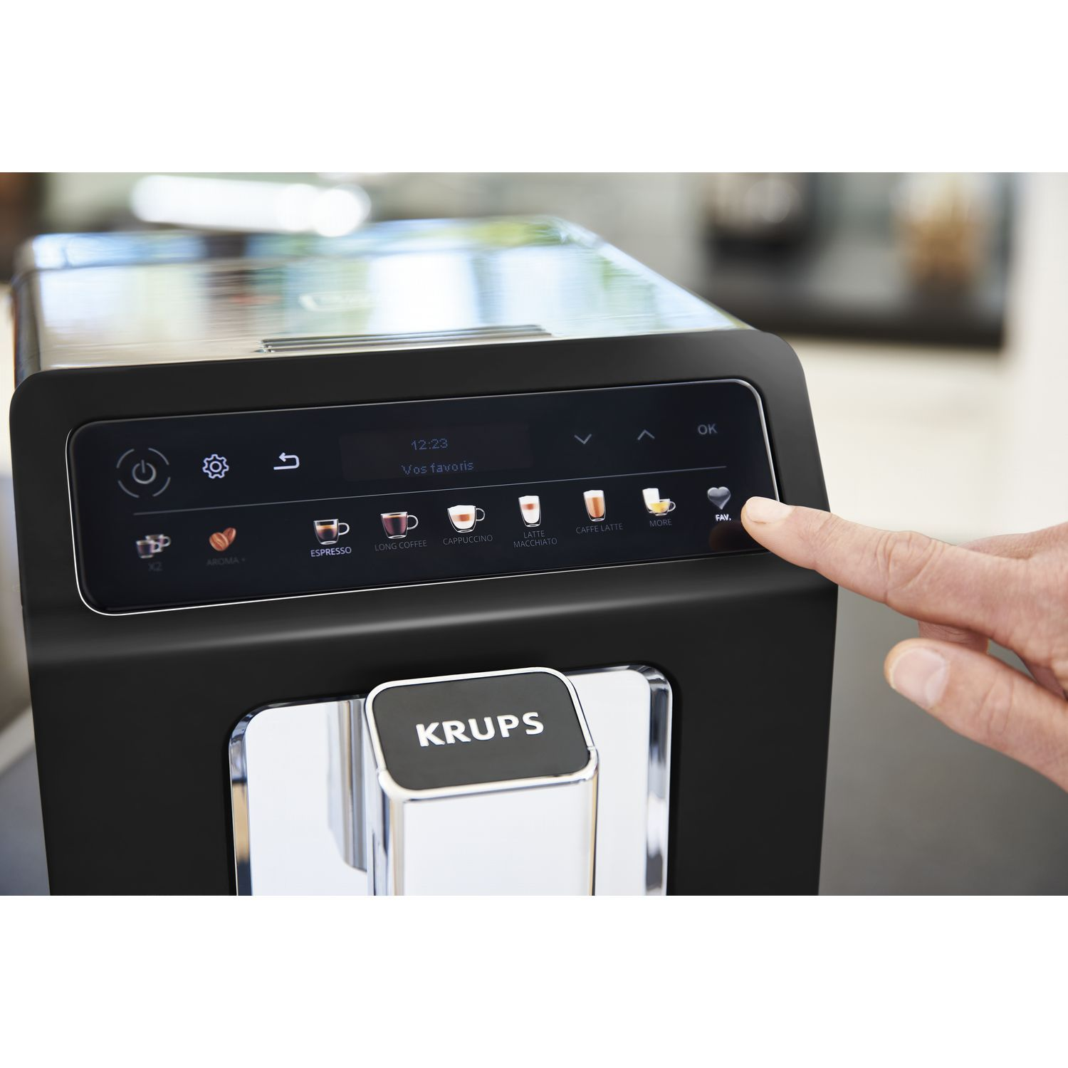 A hand presses a button on a black Krups coffee machine. The screen displays coffee options.