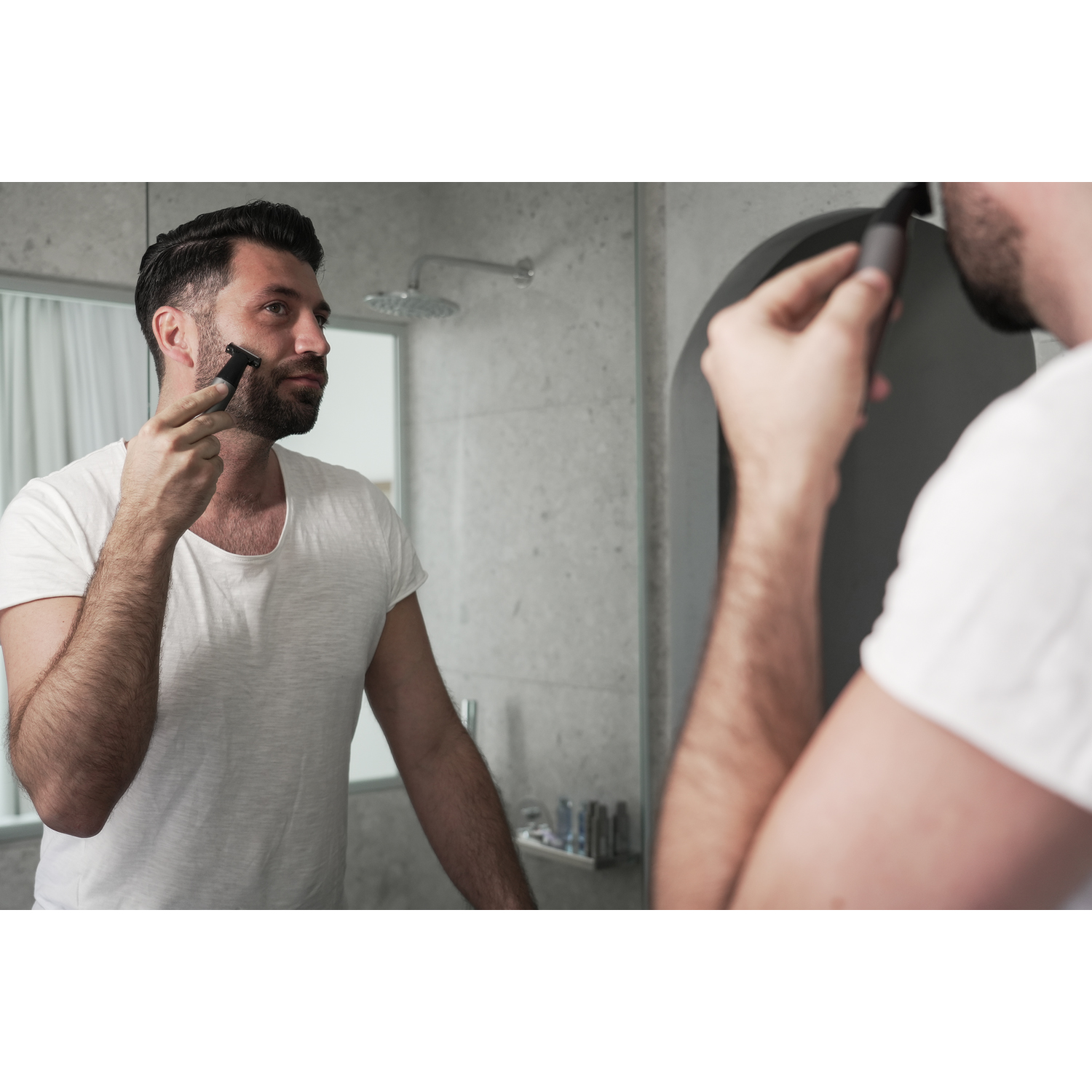 Uomo che si rade, nello specchio, maglietta bianca, ambiente bagno, che tiene il trimmer.