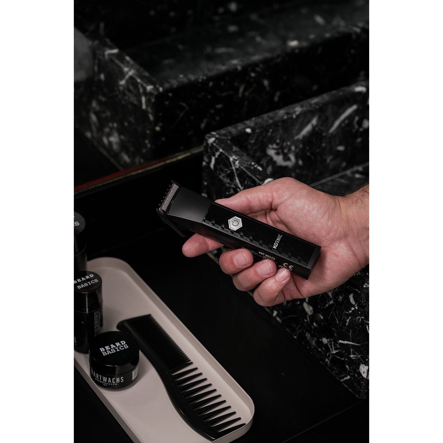 Una mano che tiene un trimmer, con prodotti per la barba e un pettine.
