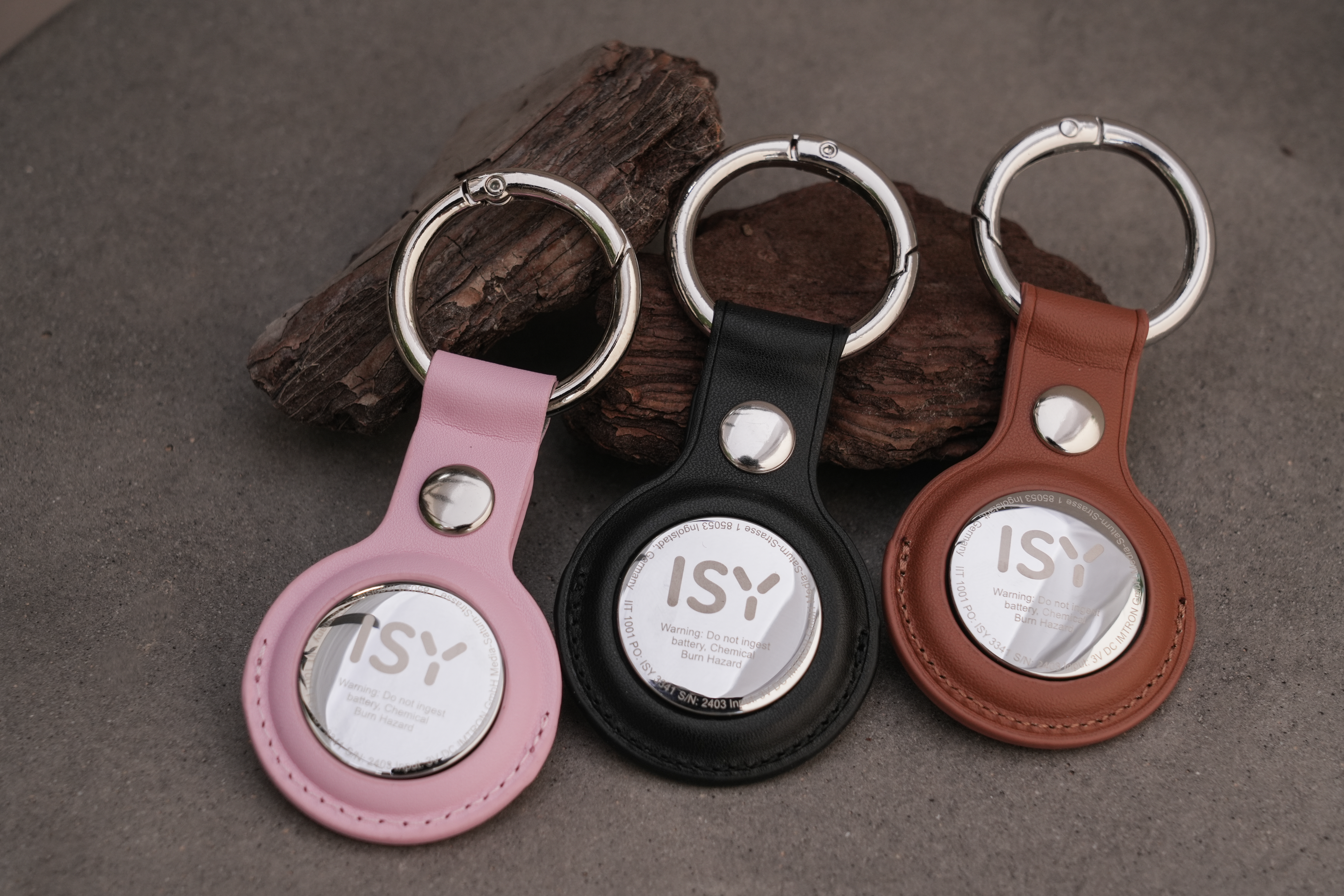 Drei Schlüsselanhänger mit dem ISY-Logo, in Rosa, Schwarz und Braun, mit silbernen Ringen, auf einer Steinoberfläche.