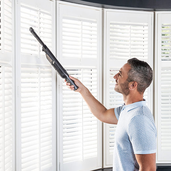 Ein Mann reinigt weiße Fensterläden mit einem schwarzen Handstaubsauger.