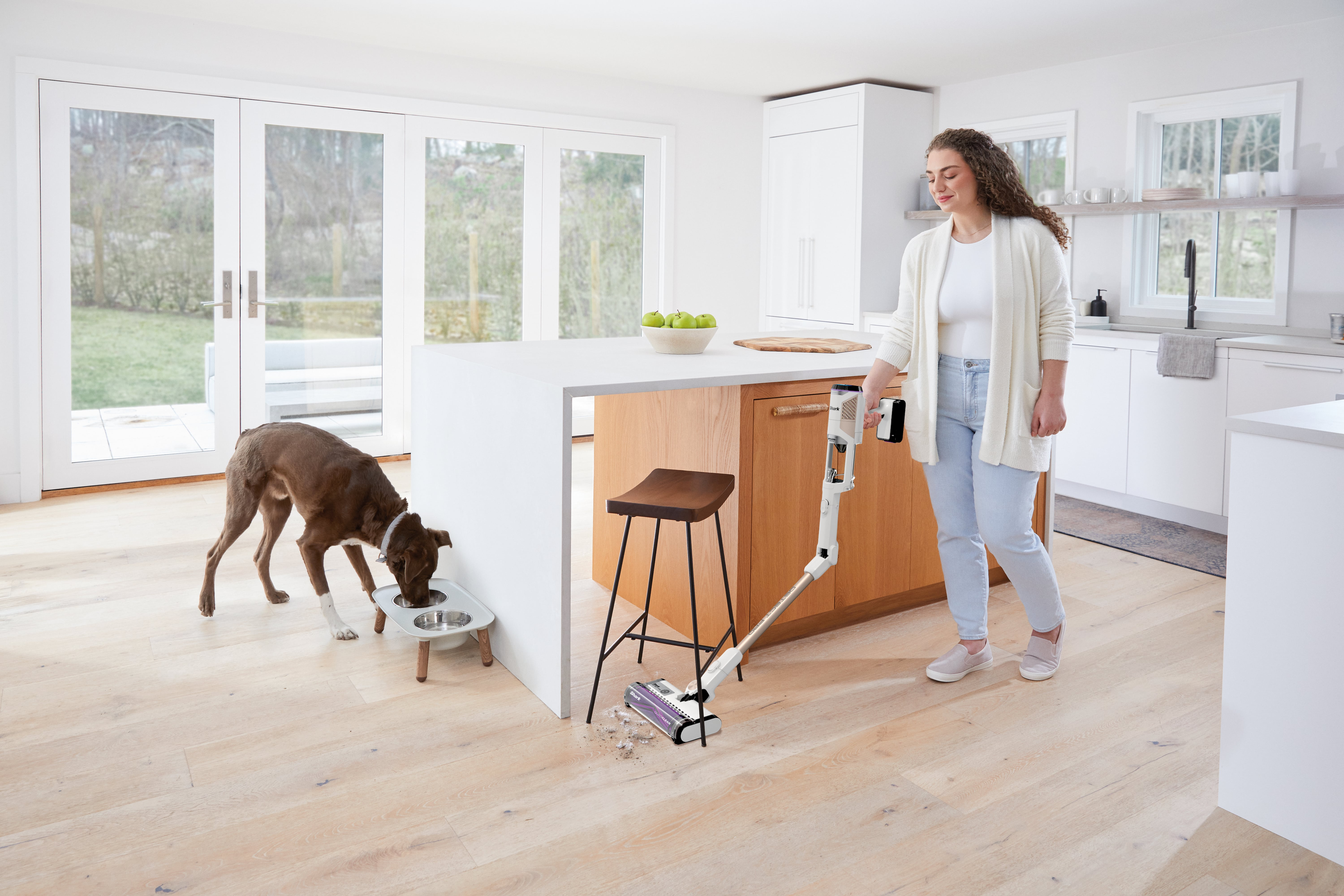 Eine Frau saugt eine Küche. Ein Hund frisst aus einer Schüssel. Die Küche ist weiß.