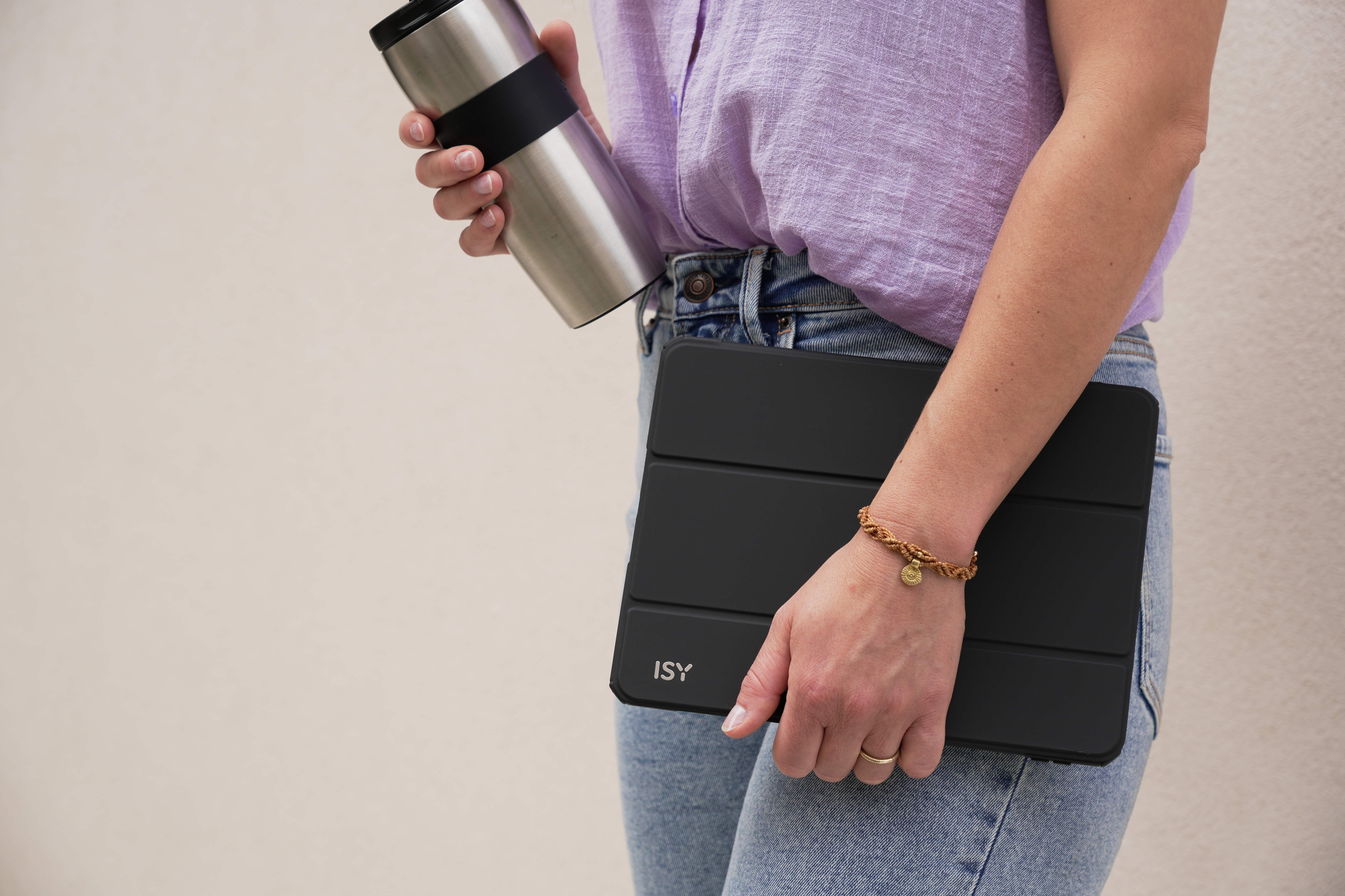 Eine Person, die eine silberne Thermoskanne und ein schwarzes Tablet hält, trägt ein lila Hemd und Jeans.