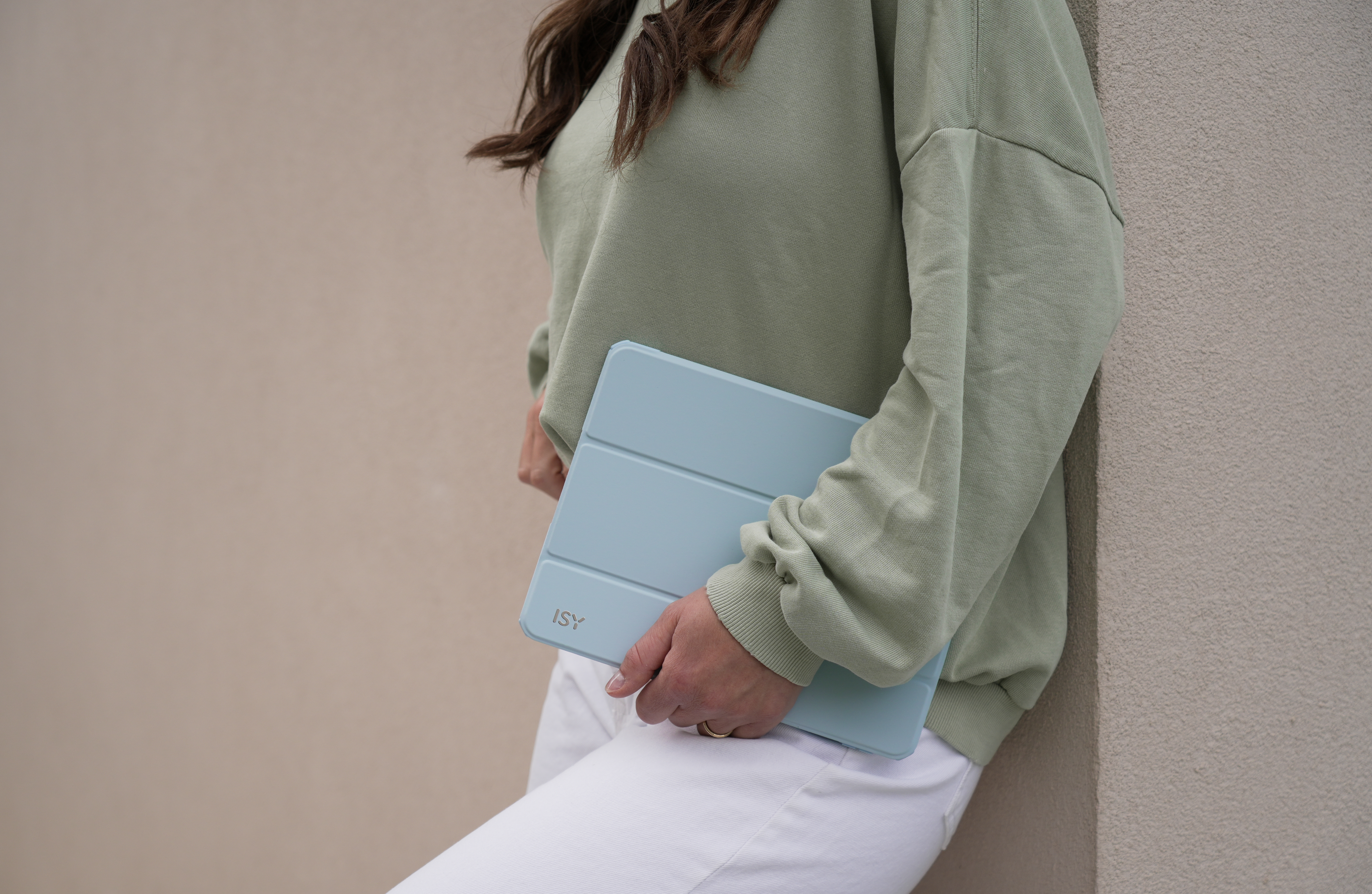 Eine Person hält eine hellblaue Tablet-Hülle und lehnt sich an eine Wand. Die Hülle hat das 'ISY'-Logo.