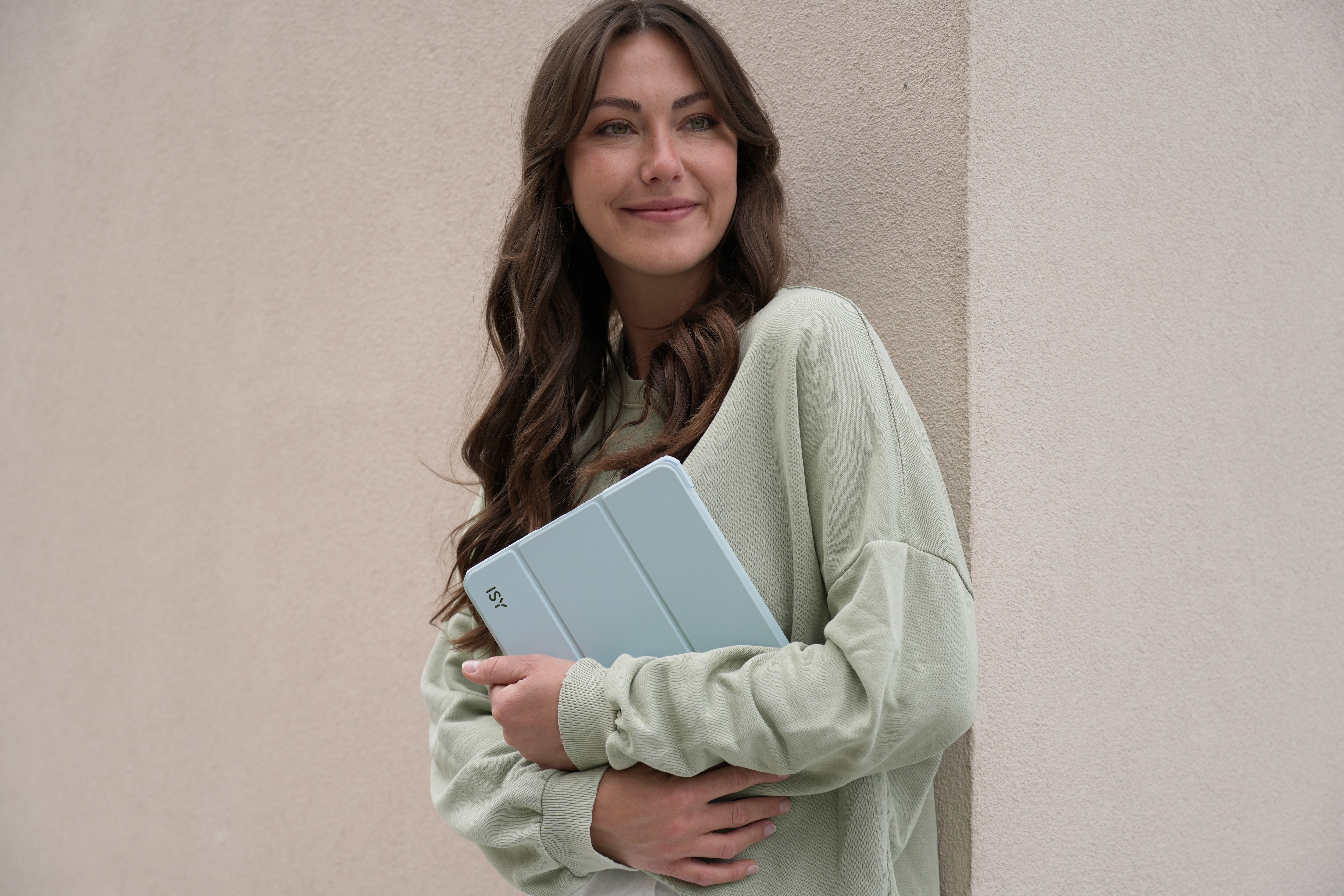 Frau mit langen braunen Haaren lächelt, hält hellblaue Tablet-Hülle neben beiger Wand.
