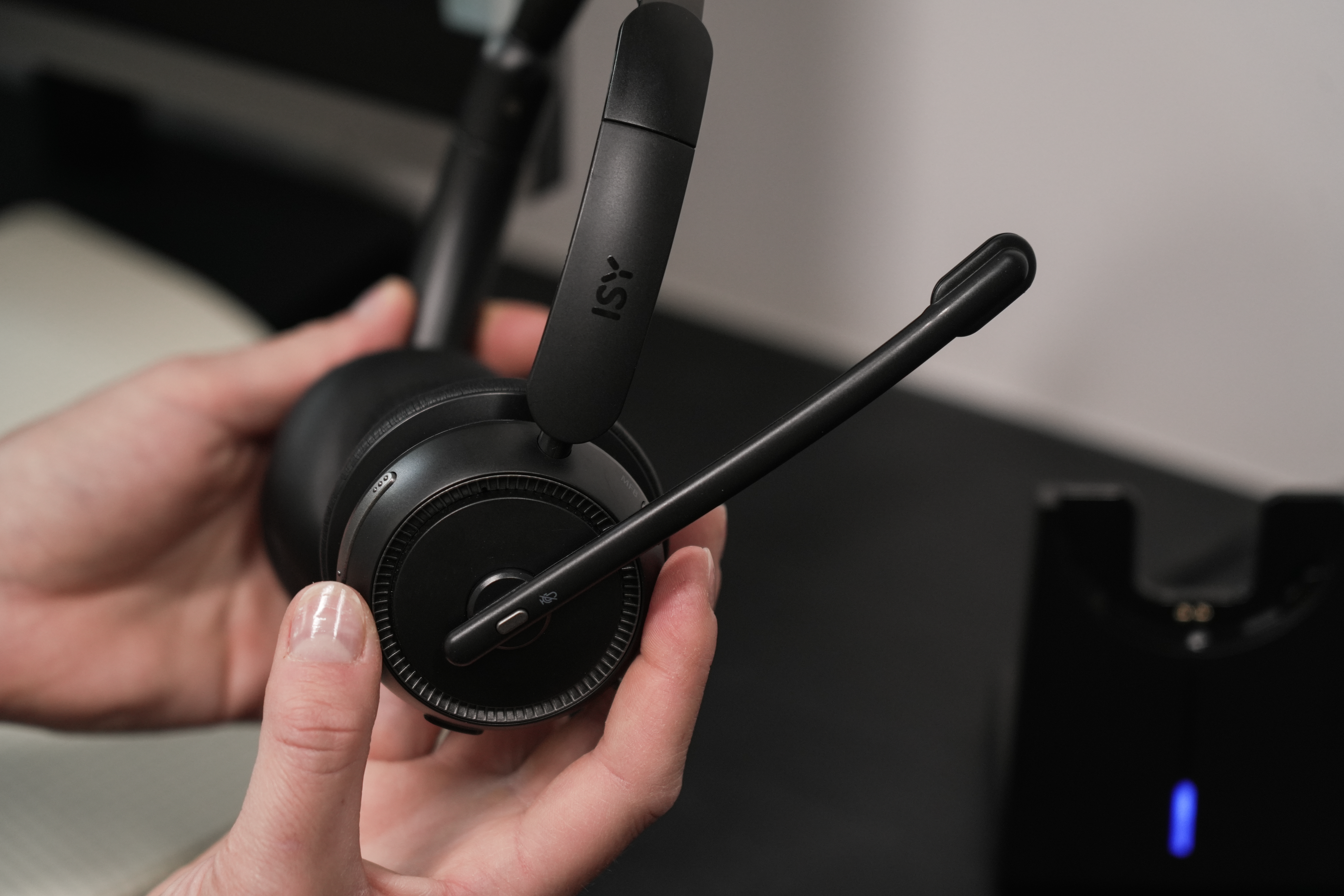 Hände halten ein schwarzes Headset mit Mikrofon und Ladestation auf dunkler Oberfläche.