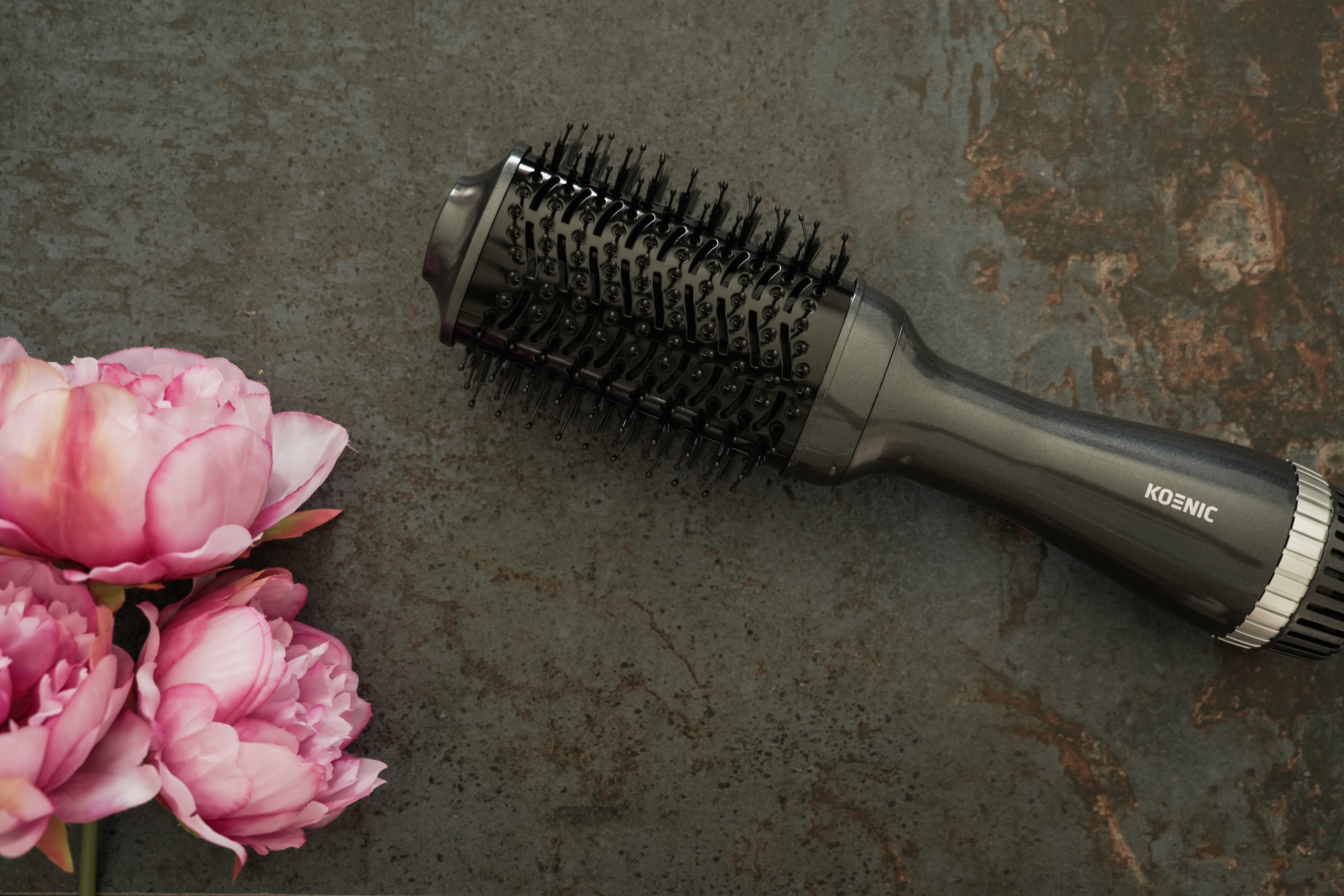 Eine schwarz-graue Haartrocknerbürste und rosa Blumen auf dunklem Hintergrund.