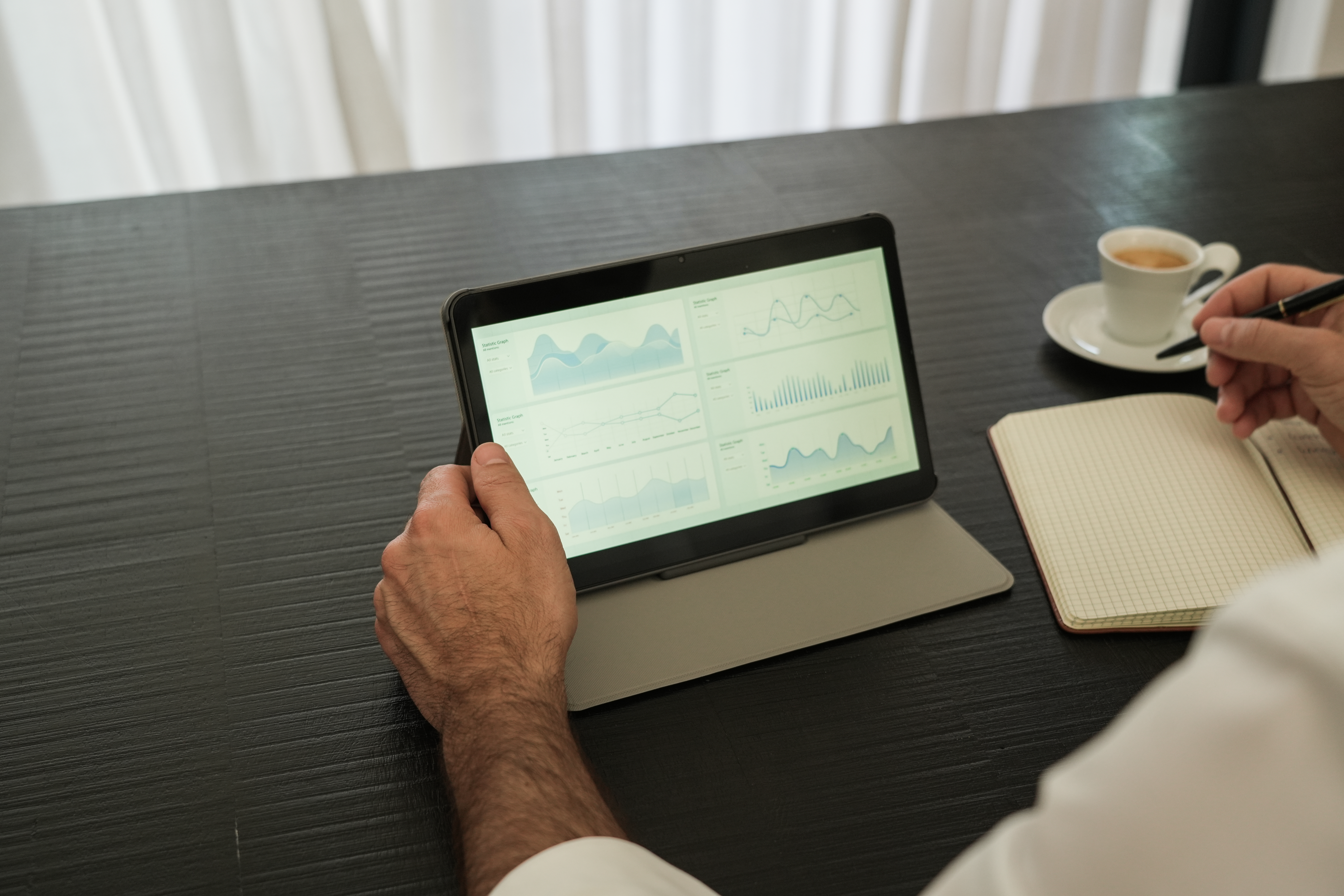 Eine Person arbeitet an einem Tablet, mit einer Tasse Kaffee und einem Notizbuch auf einem Tisch.