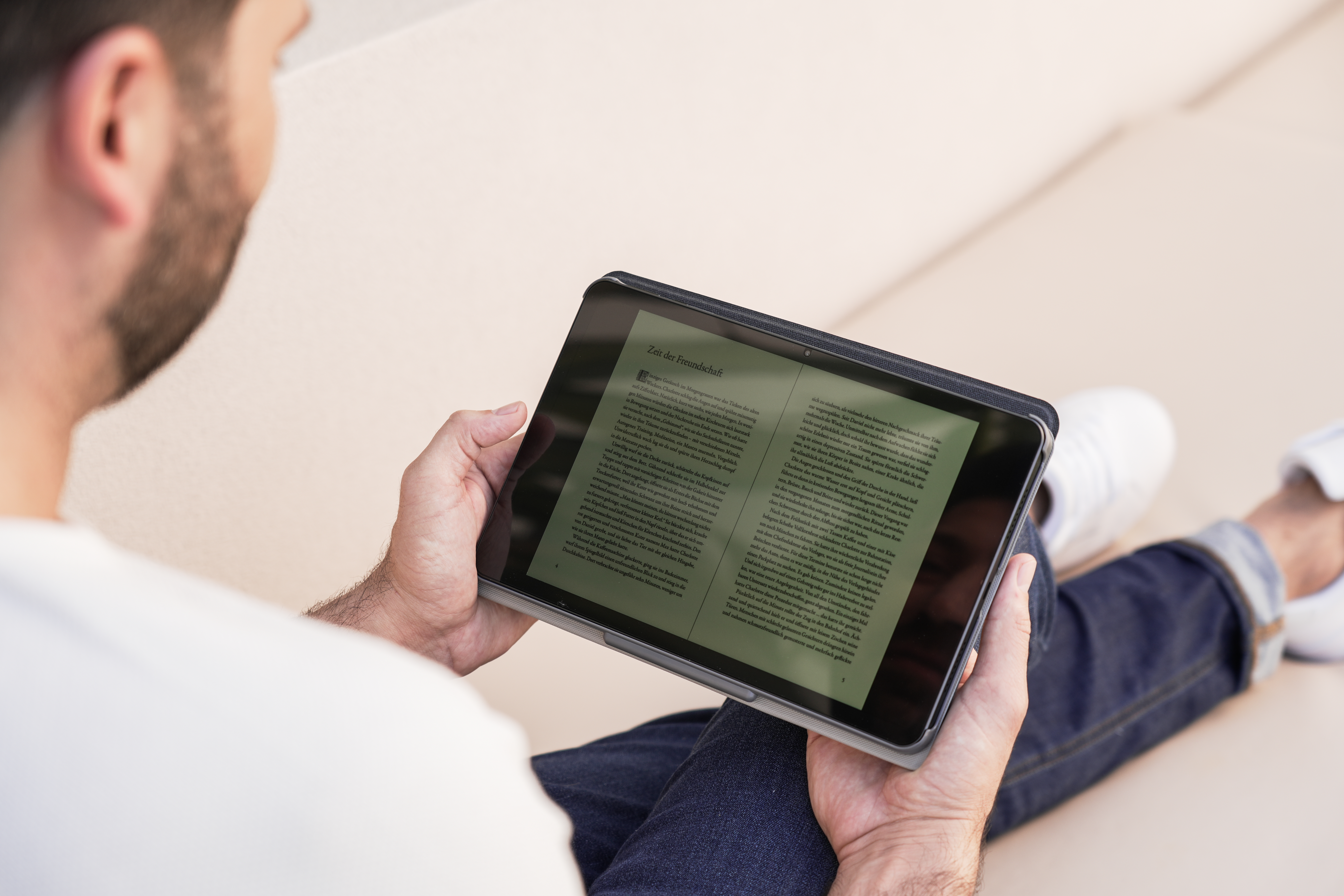 Eine Person liest auf einem Tablet, liegt auf einem Sofa, Hände halten das Gerät.