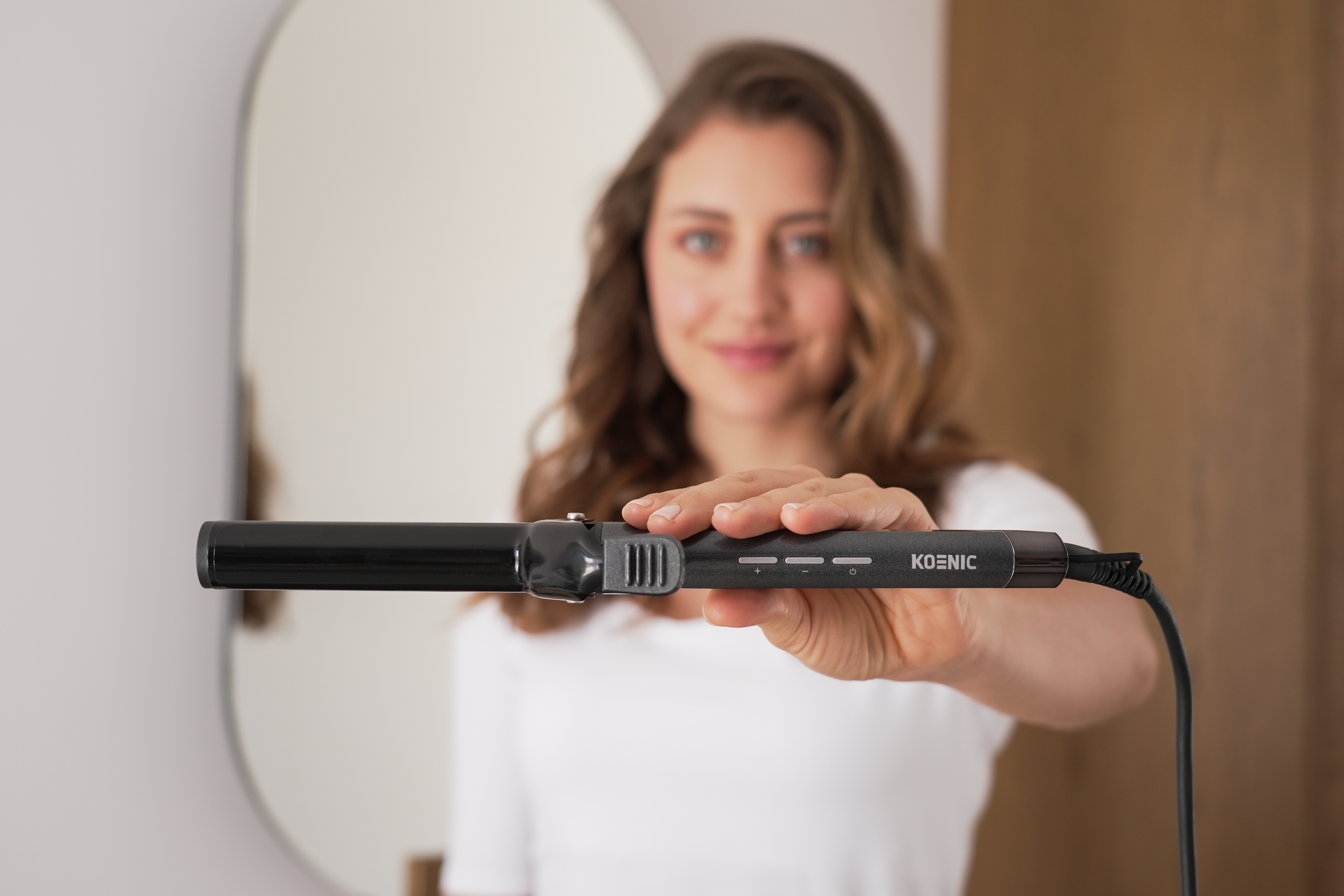 Eine Frau hält einen Koenic Lockenstab, lächelt, mit einem Spiegel im Hintergrund.
