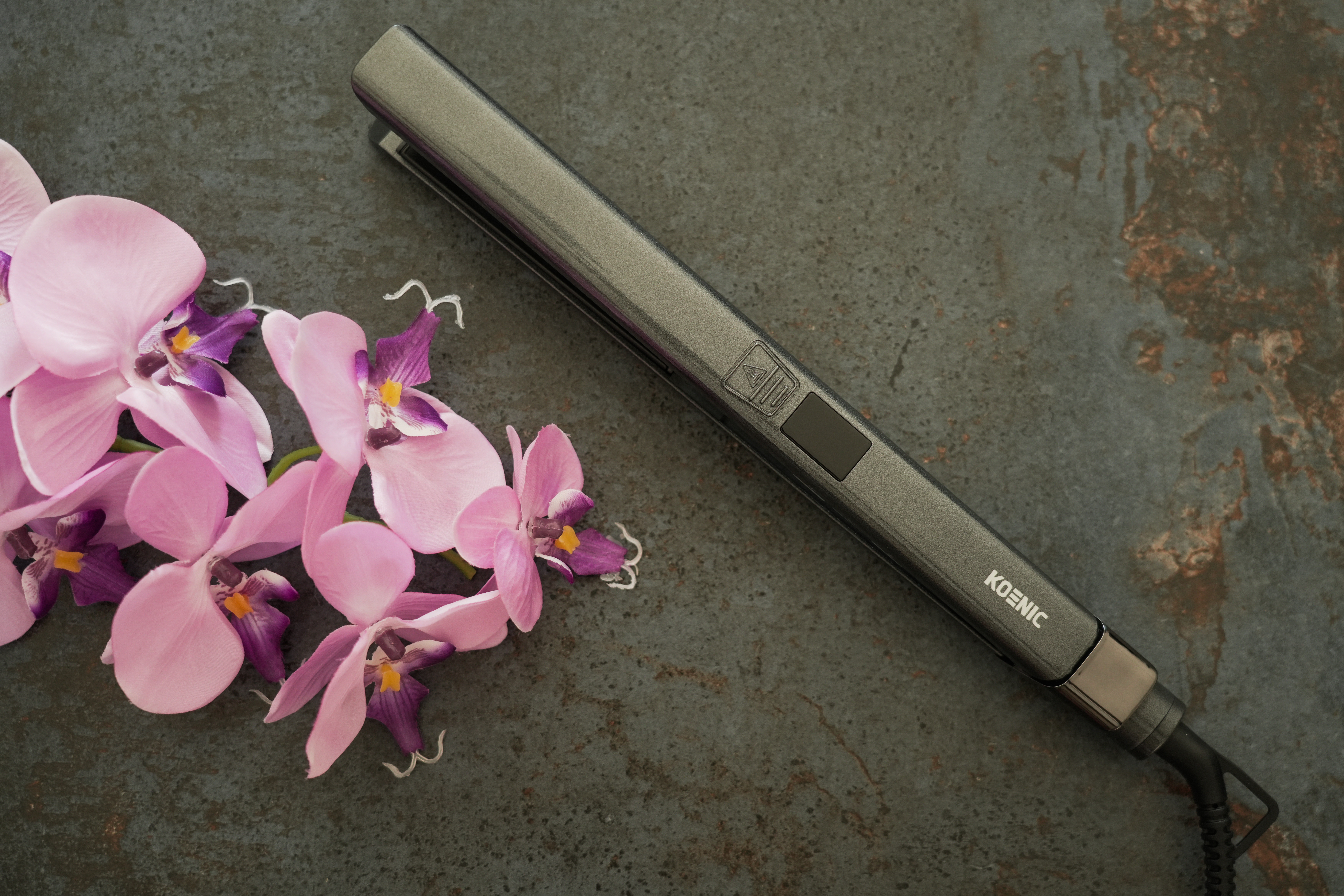 Ein grauer Haarglätter und rosa Orchideen auf einer dunklen, strukturierten Oberfläche.
