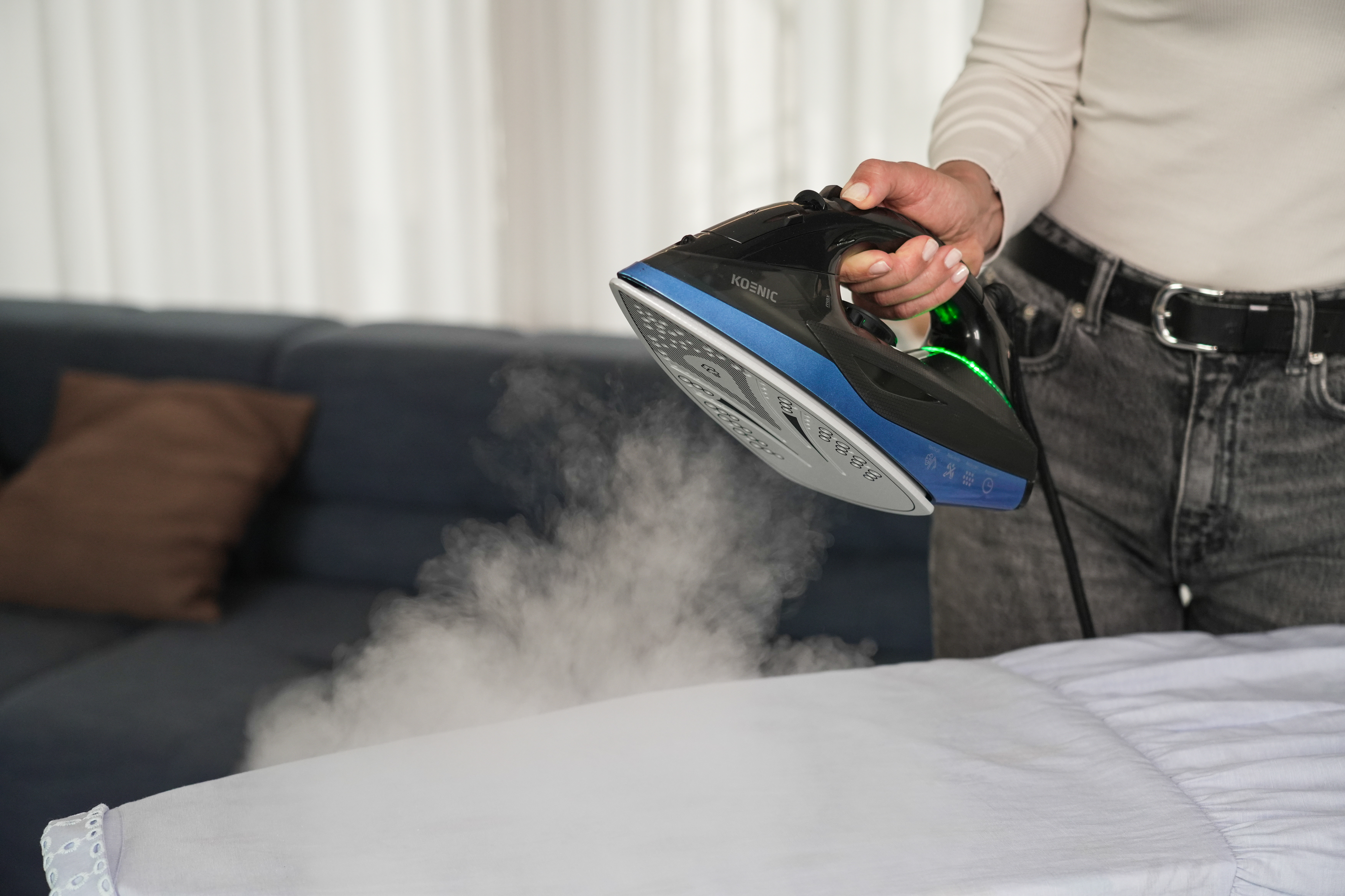 Person bügelt weißes Hemd mit Dampf, mit einem schwarz-blauen Bügeleisen auf blauem Sofa.