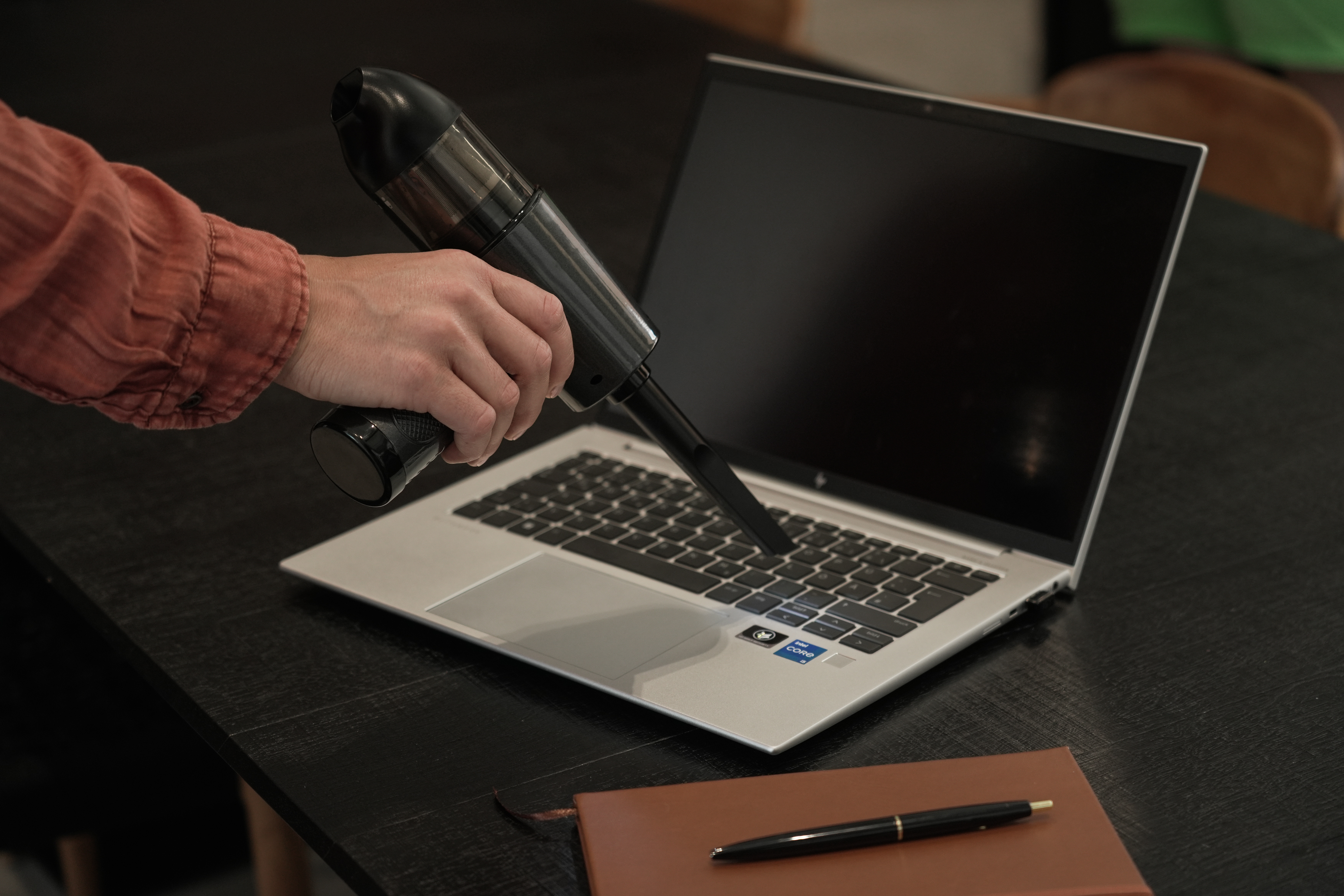 Eine Person reinigt einen Laptop mit einem Staubsauger auf einem schwarzen Tisch. Ein Notizbuch und Stift liegen auch auf dem Tisch.