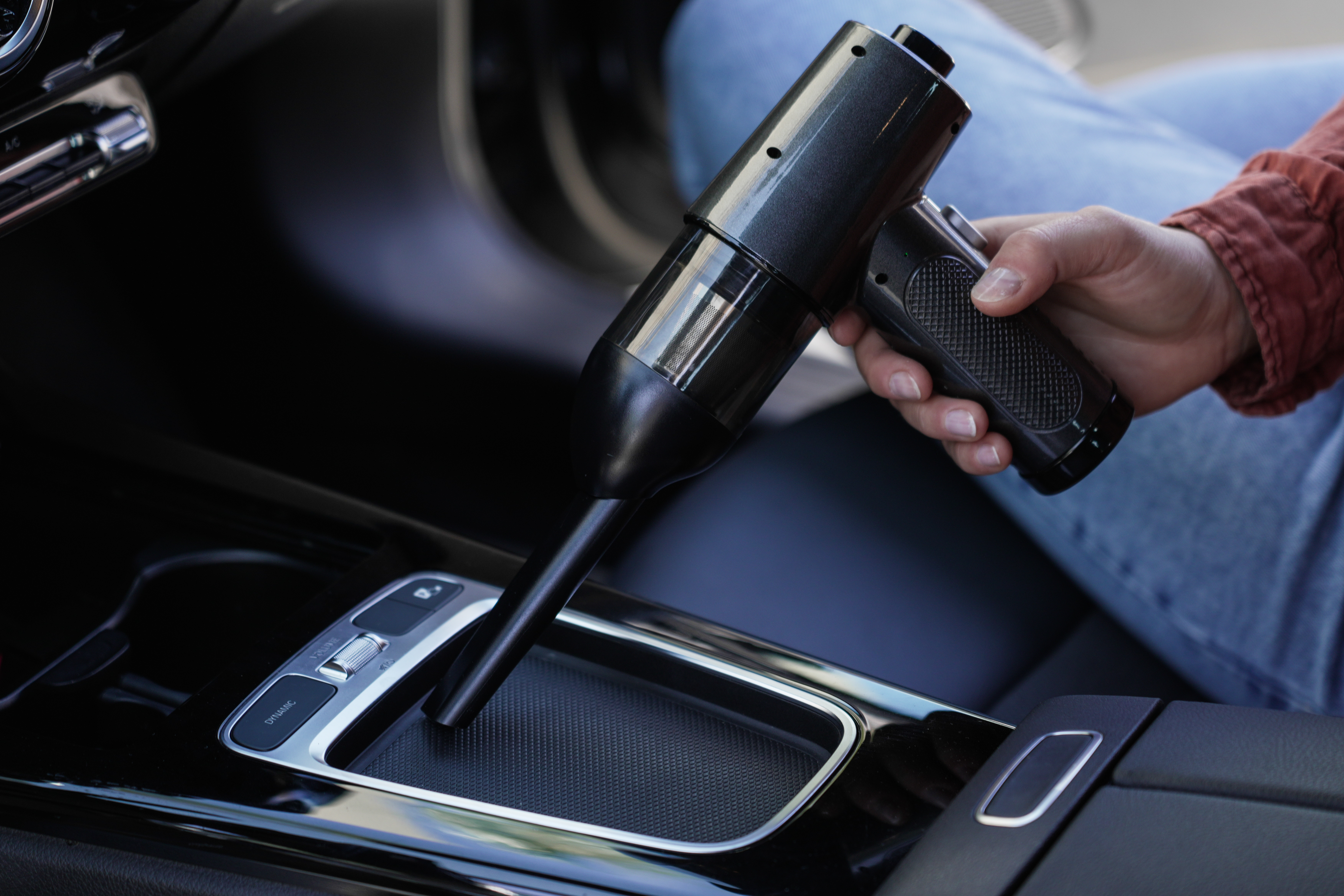 Hand hält einen schwarzen Staubsauger in einem Auto. Er soll die Mittelkonsole des Autos reinigen.