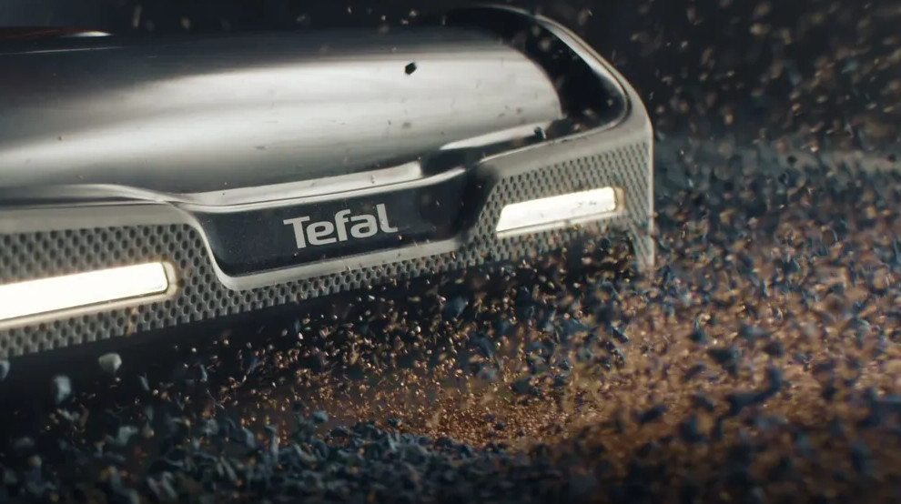 Zbliżenie urządzenia Tefal z logo, z unoszącymi się odłamkami. Ciemne tło.