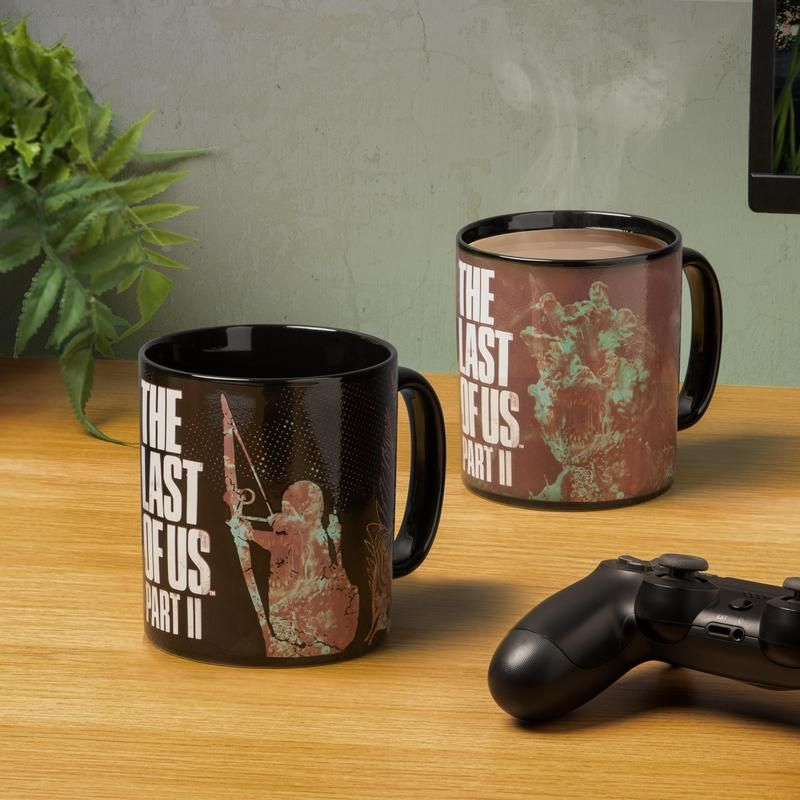 Két fekete bögre a 'The Last of Us Part II' logóval, egy kontrollerrel és egy növénnyel egy fa asztalon.